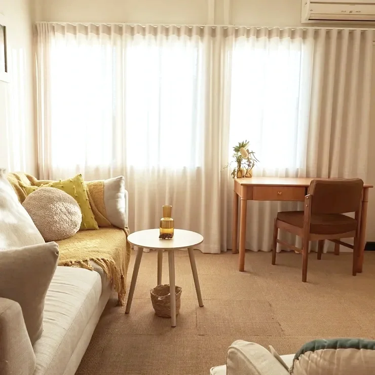Bright living room with large window covered by cream curtains, beige sofa with various pillows, small white coffee table with a bottle, and a wooden desk with a chair and a plant in a vase.