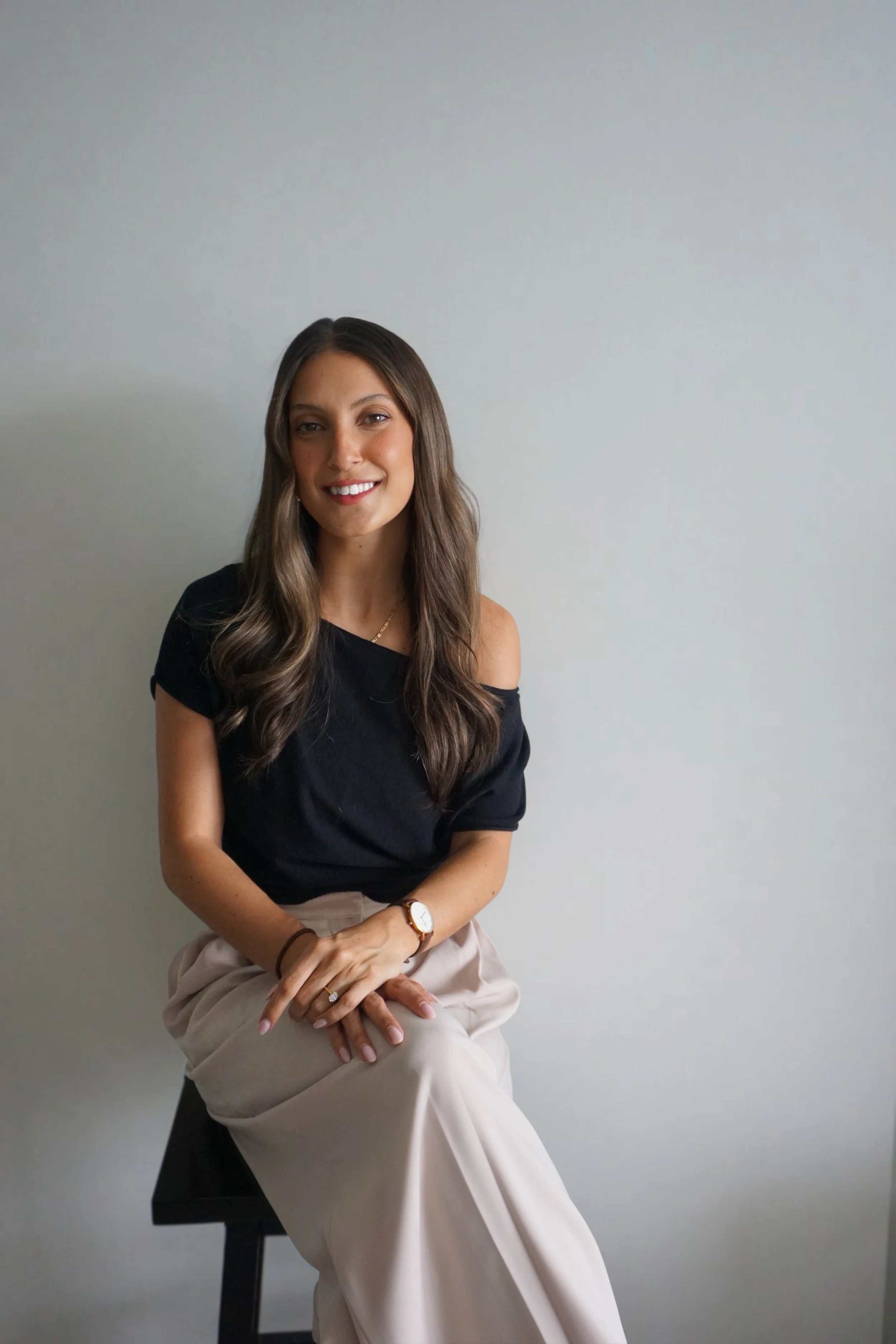 A woman with long brown hair wearing a black top with shoulder cutouts and beige pants, sitting on a black stool against a plain light-colored wall, smiling at the camera.