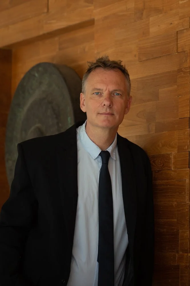 A man in a black suit and white shirt with a black tie standing against a wooden wall, looking at the camera.