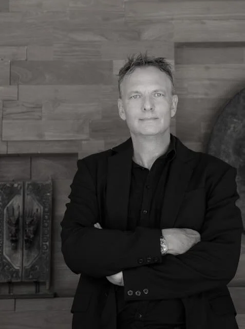 A man in a black suit with arms crossed, standing against a wooden wall with artwork.