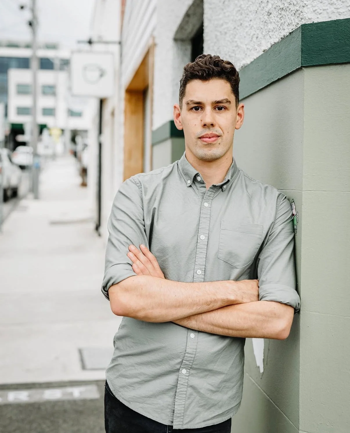 Chef Jono, a young man with crossed arms leaning against a wall on a city sidewalk, wearing a grey button-up shirt, with blurred buildings and street in background.