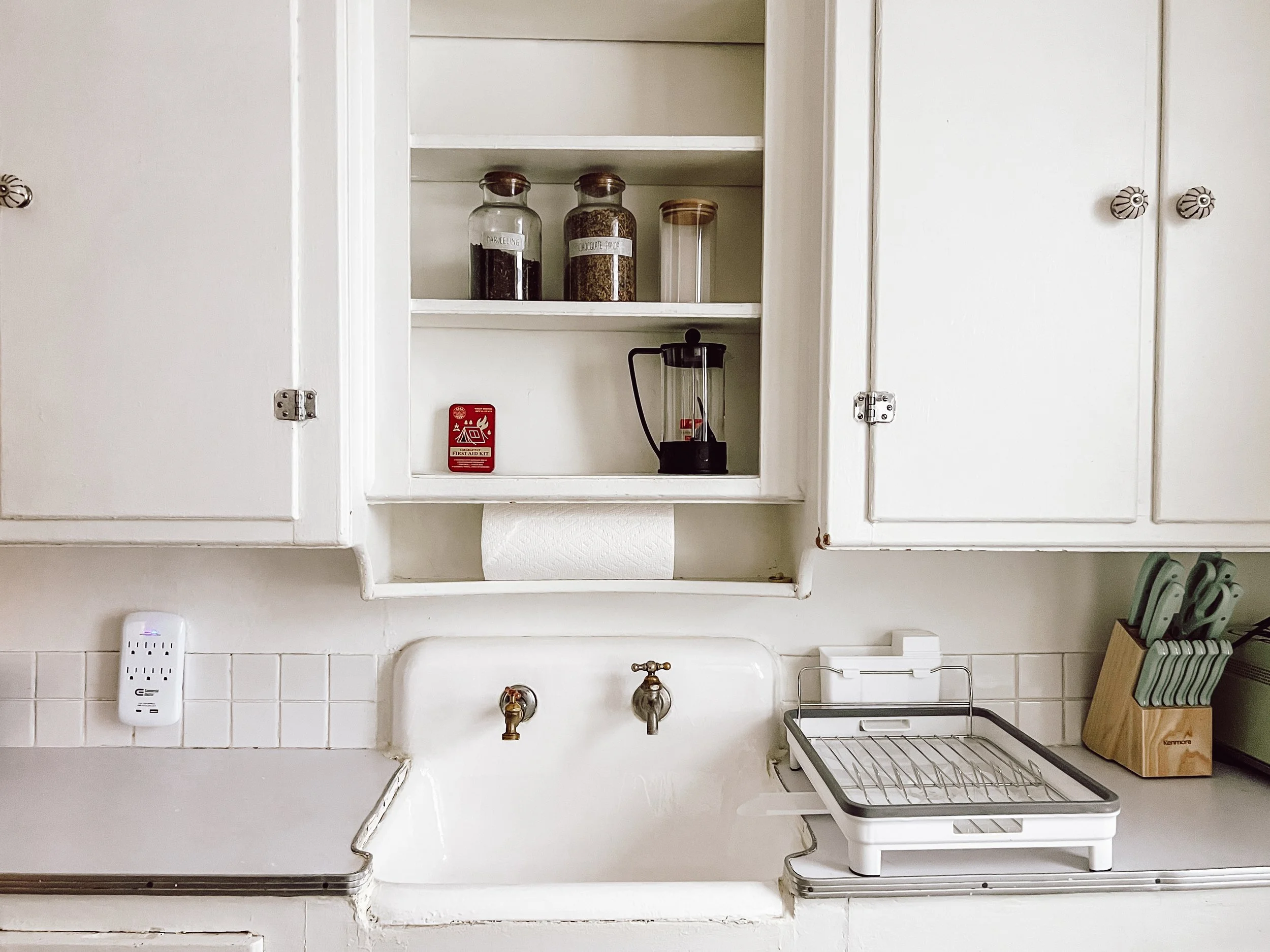 Kitchen with white cabinets, open shelves holding jars of spices, a lantern, and a red fire alarm. White sink with brass faucet, gray dish rack, and a knife block with green-handled knives on the countertop.
