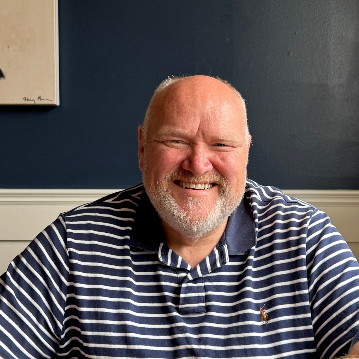 A smiling, bald, bearded man with light skin wearing a navy blue and white striped polo shirt, sitting indoors against a dark blue wall.