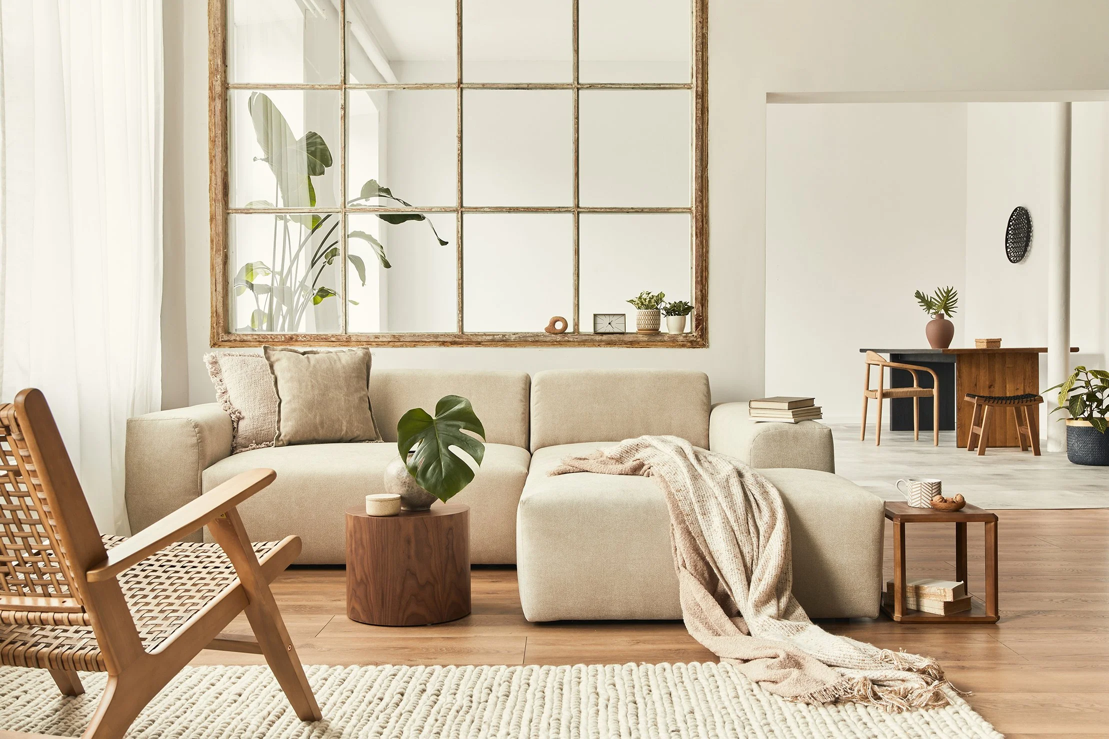 Bright and airy living room with beige sectional sofa, wooden side table, and woven armchair. Large window with traditional grid panes, lush green plants, and minimalistic decor create a cozy, modern atmosphere.