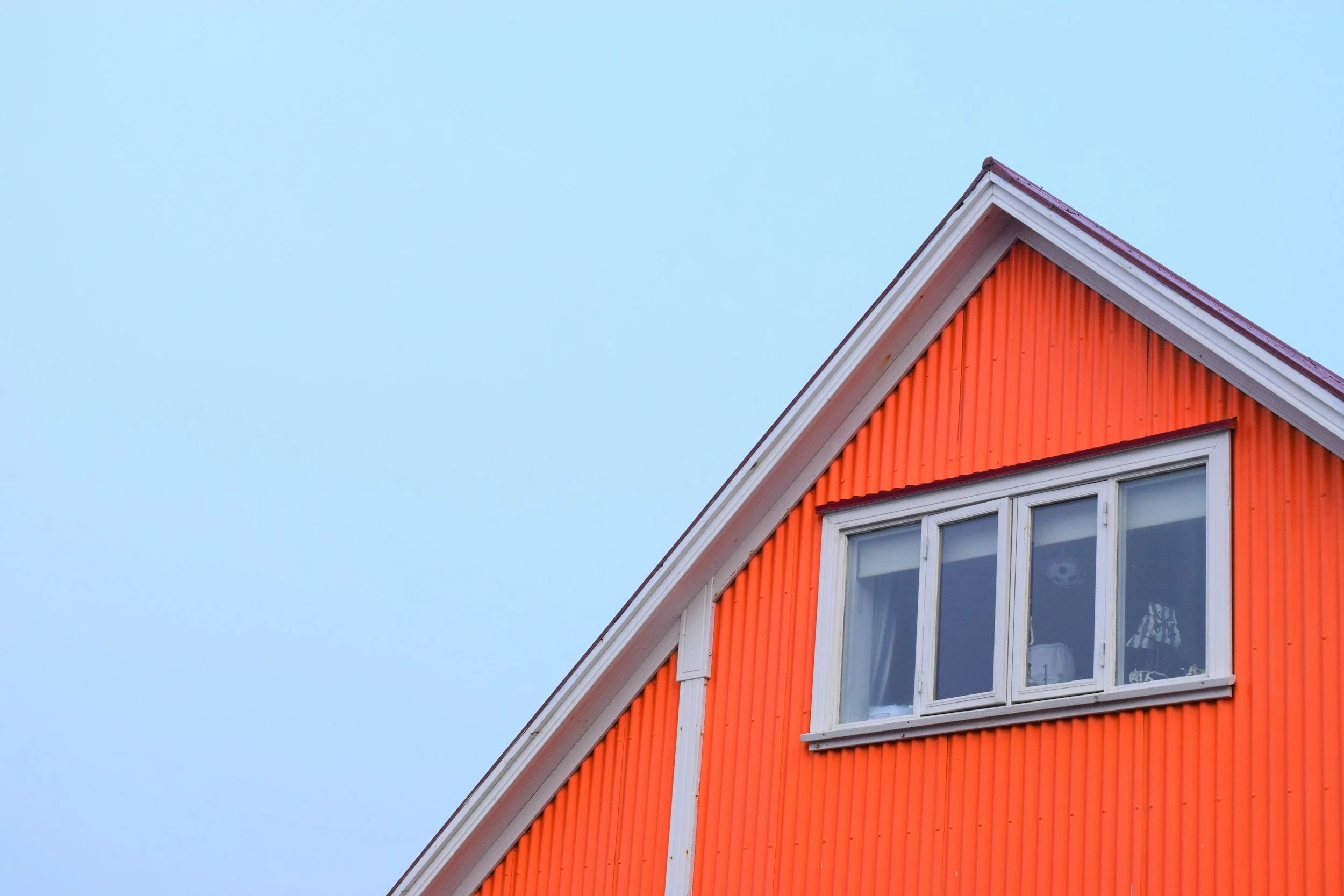 Close-up of the orange exterior wall and white window of a house with a peaked roof against a clear blue sky.