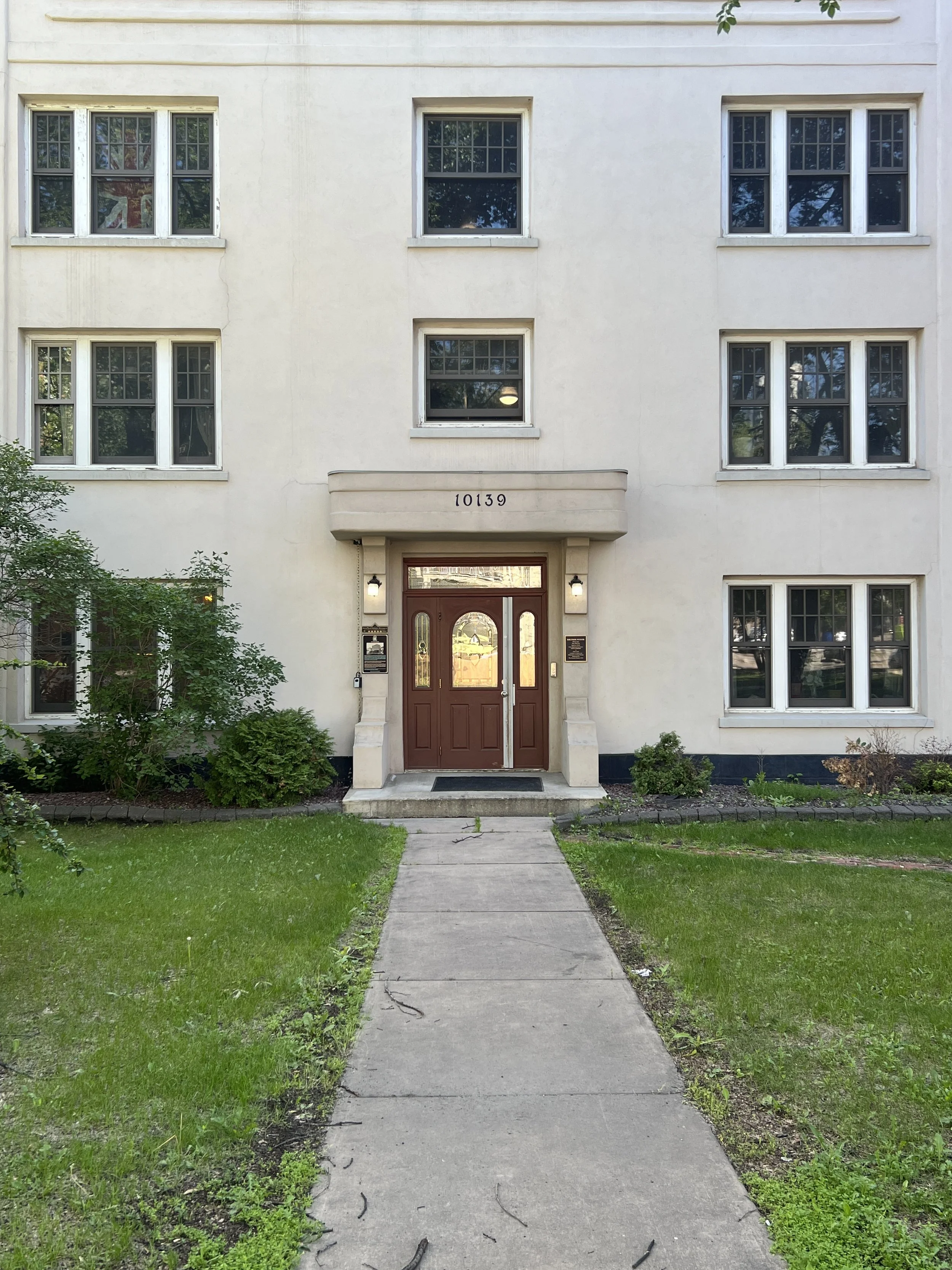 Front entrance of a white multi-story residential building with a brown door, the number 10139 above it, and a concrete walkway leading to the door, flanked by greenery.