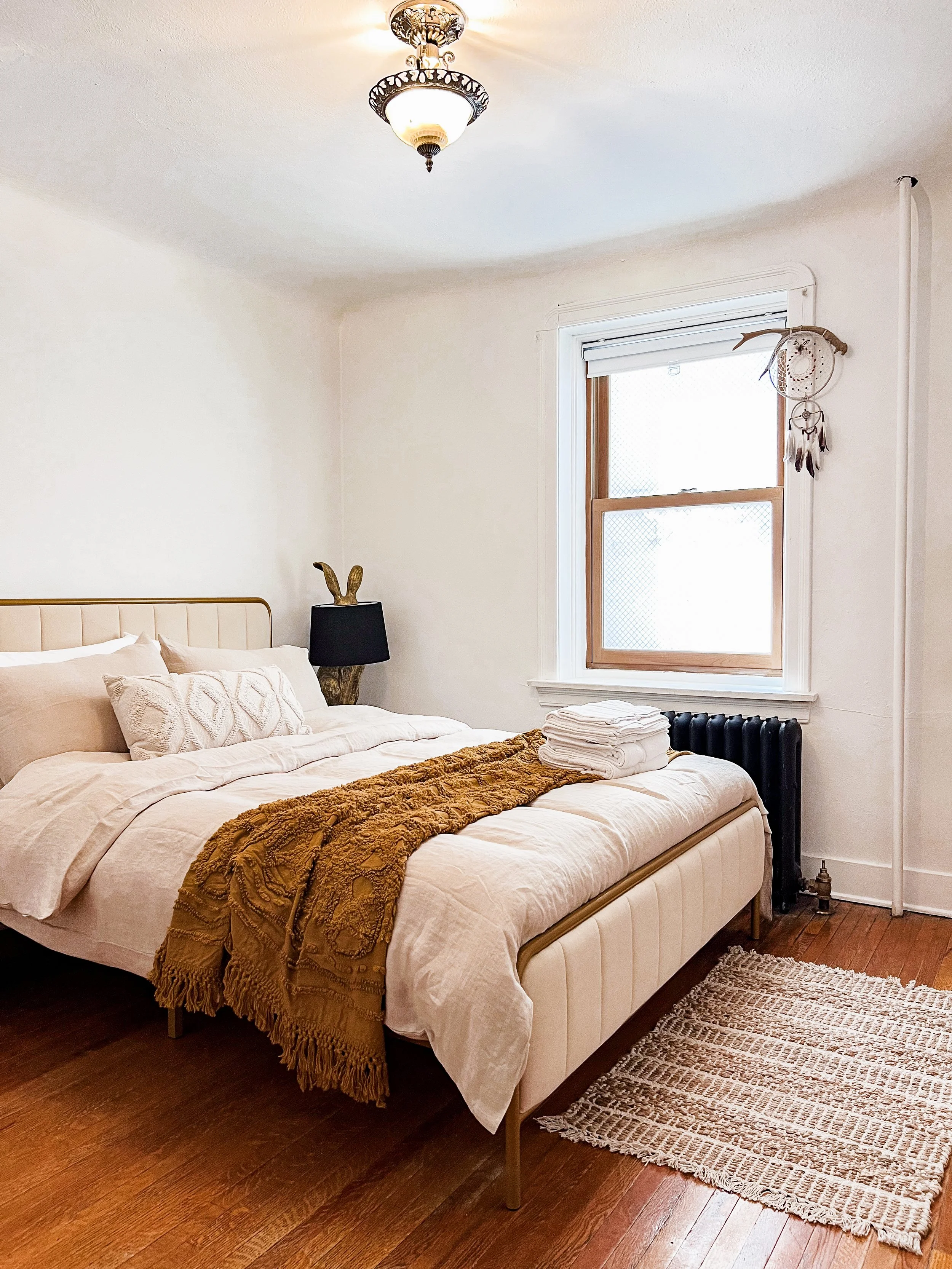 A cozy bedroom with a beige bed, white bedding, and a mustard throw blanket. There is a black lamp with a decorative base on a nightstand, a window with white blinds, and a dreamcatcher hanging near the window. A small striped rug is on the wooden floor, and folded towels are on the bed.
