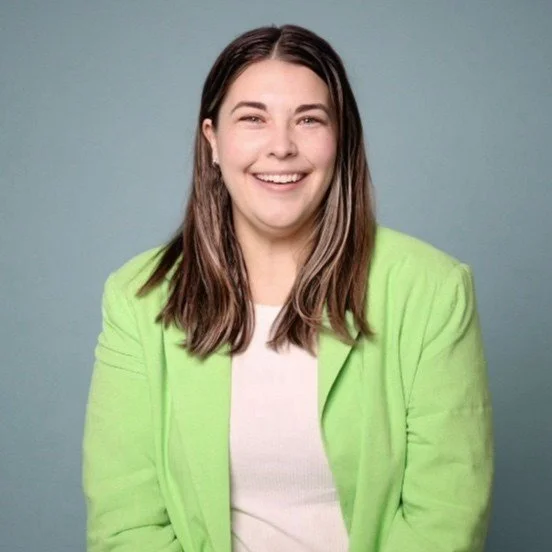 A woman with long brown hair wearing a lime green blazer and a white top, smiling and looking at the camera against a plain gray background.