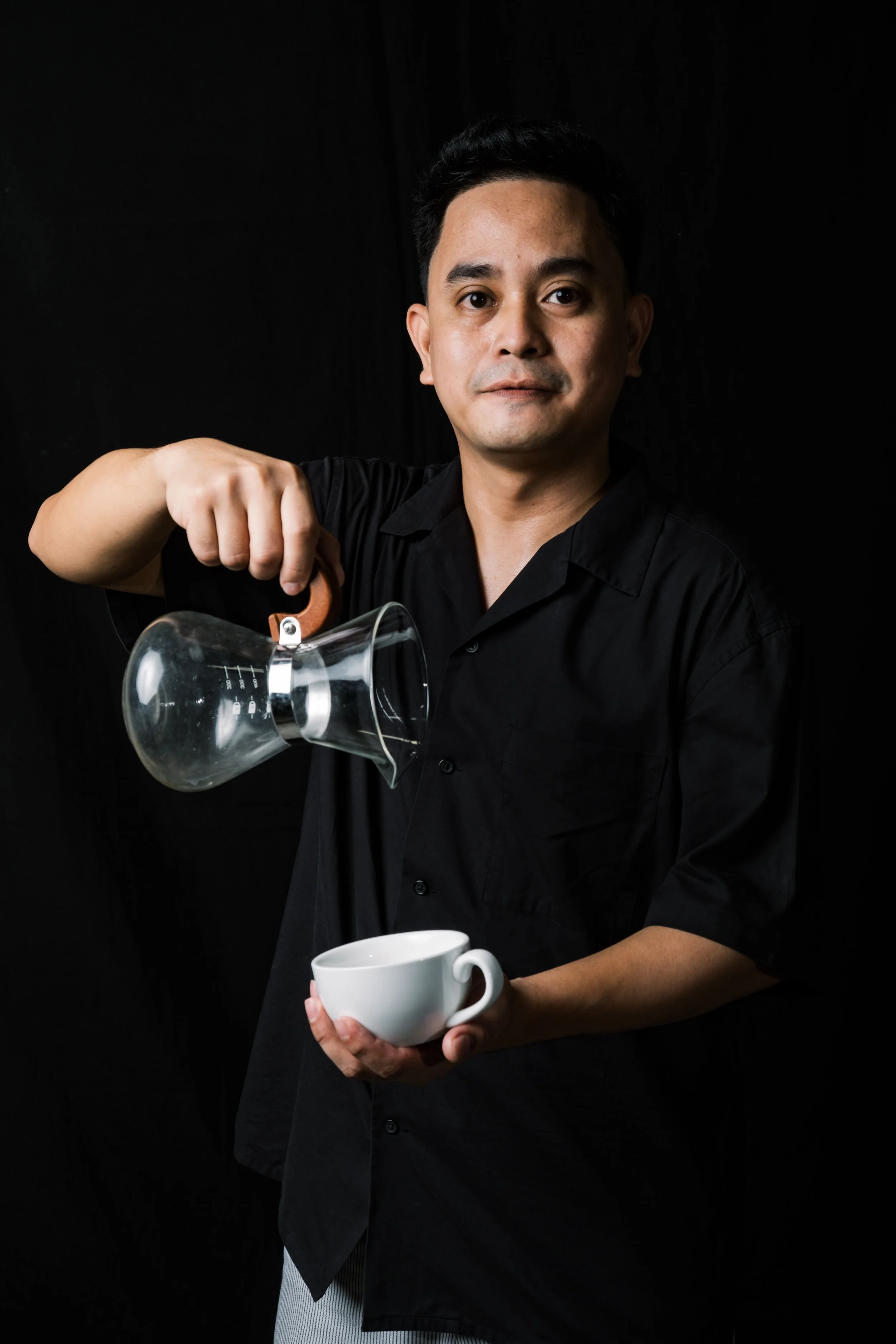 Eugene Dellagas - Man in black shirt pouring coffee into a white cup against dark background.