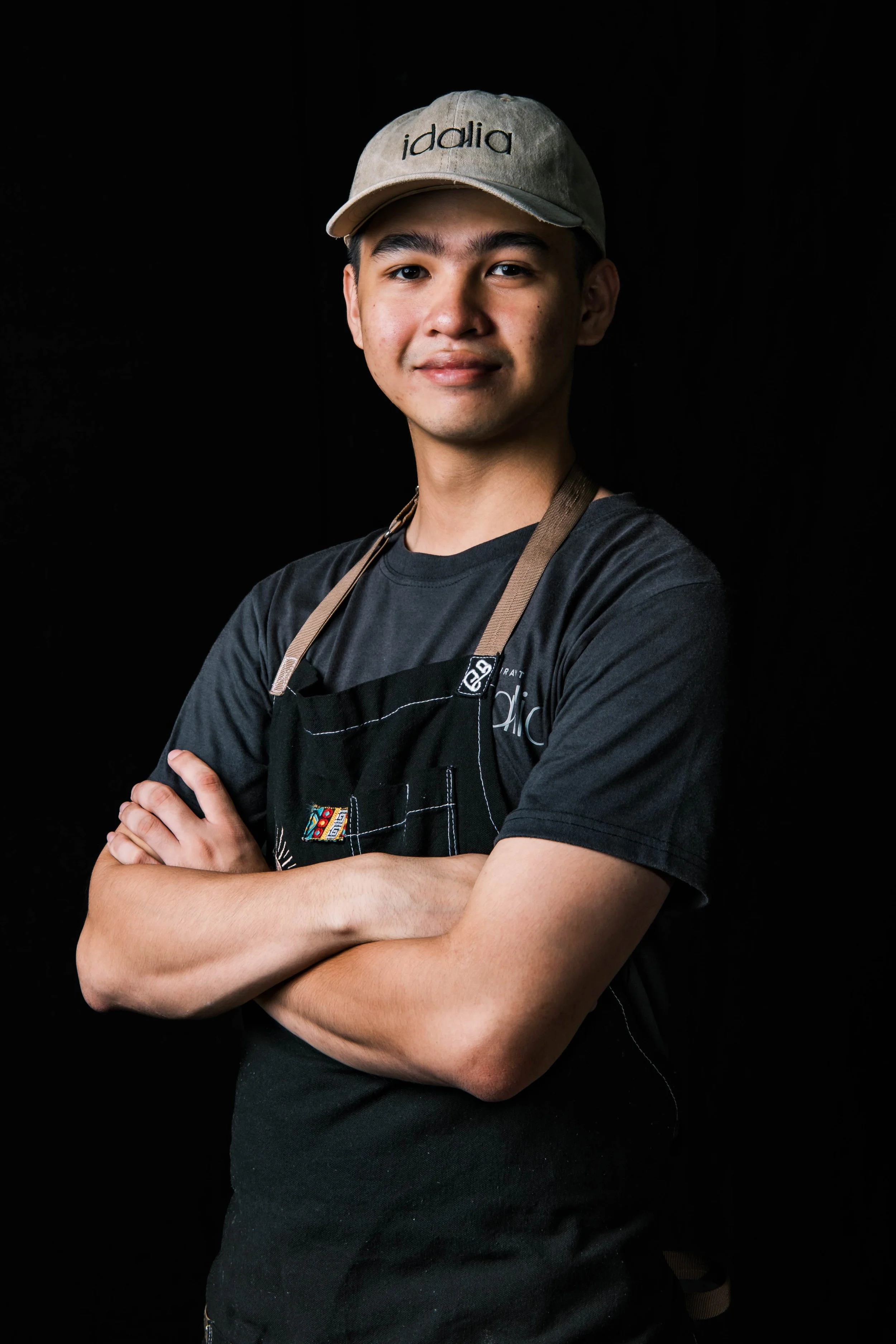 Dre Aganinta - A young man wearing a beige cap, black t-shirt, and an apron, standing against a black background with arms crossed.