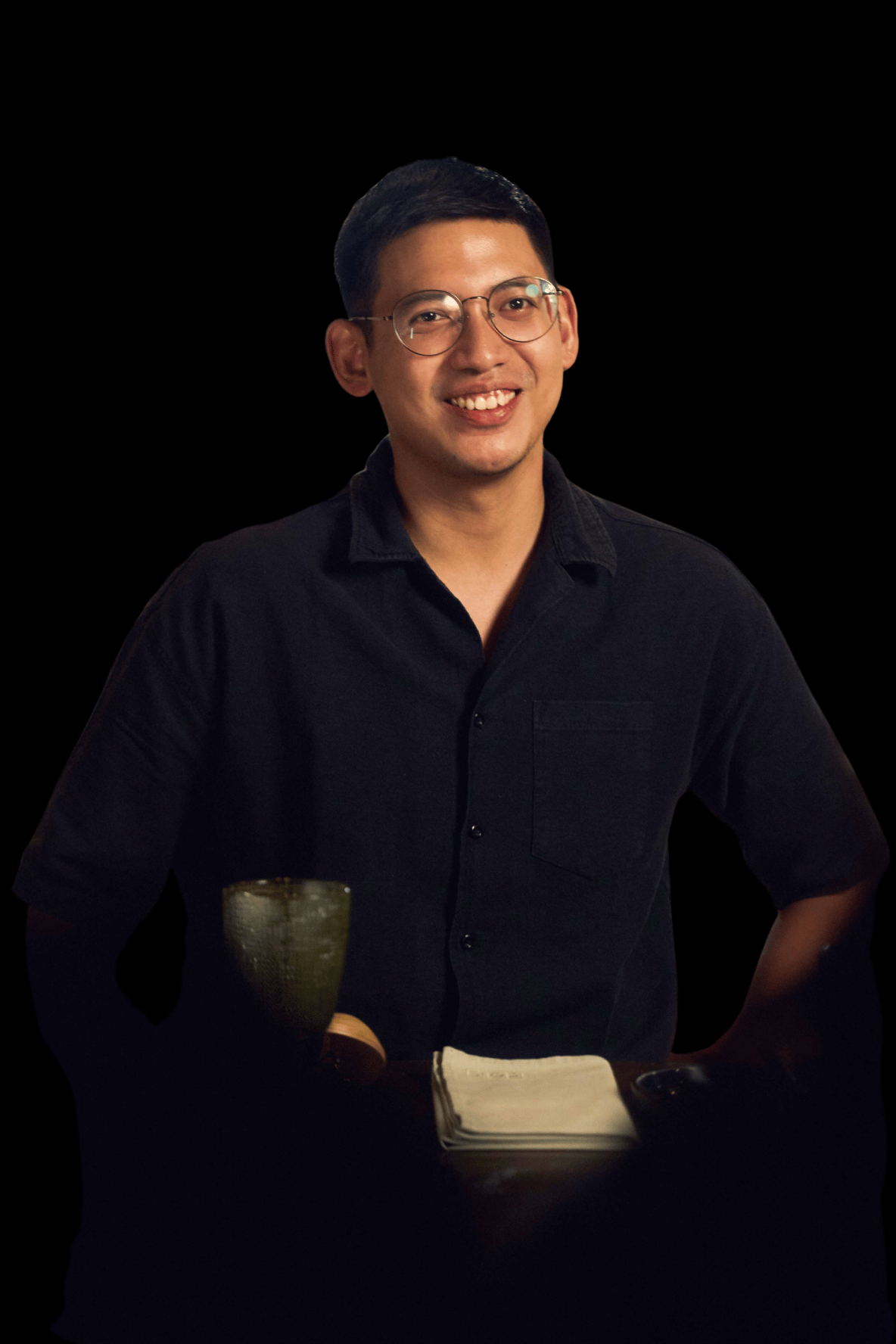 Lorenzo David - A young man with short dark hair and glasses, wearing a black button-up shirt, smiling and standing with hands on hips, in a dark environment with a notebook and a ceramic cup on the table in front of him.