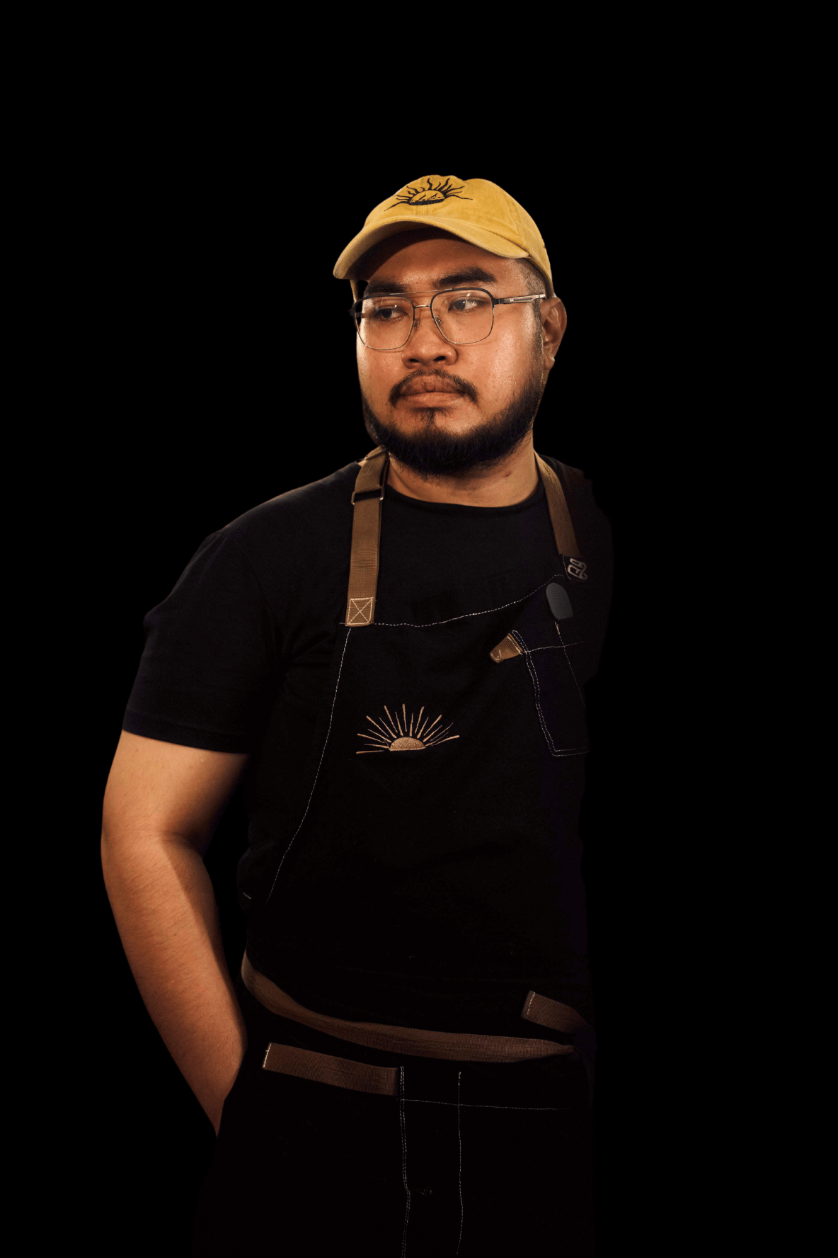 Derrick Tabu - A man with glasses and a beard, wearing a beige cap with an eye design, a black t-shirt, and an apron with a sunrise graphic, against a black background.
