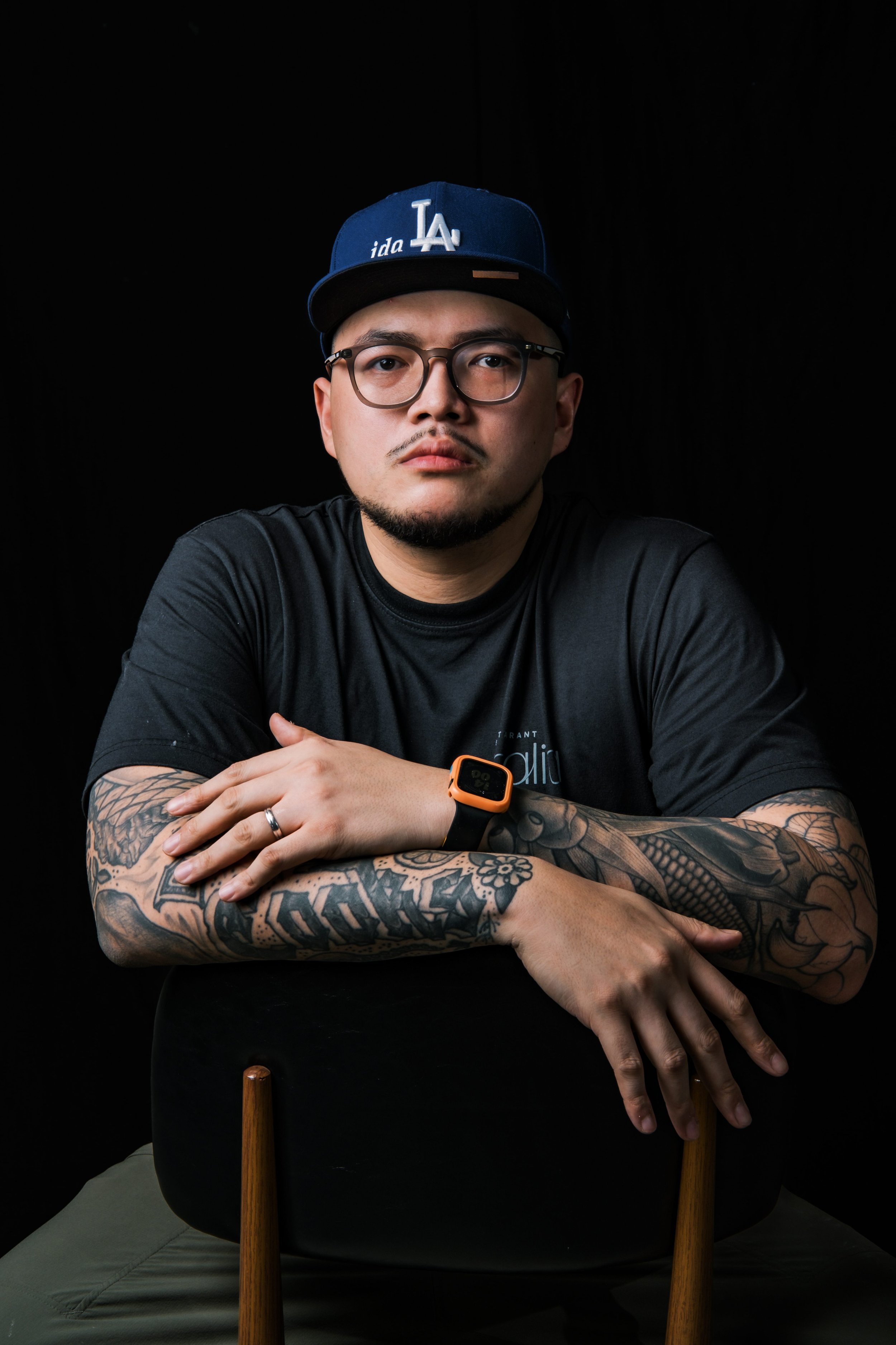 Chef Kevin David - A young man with glasses, tattoos, and a black shirt sitting behind a wooden chair with crossed arms, against a black background, wearing a blue Los Angeles Dodgers cap, with a serious expression.
