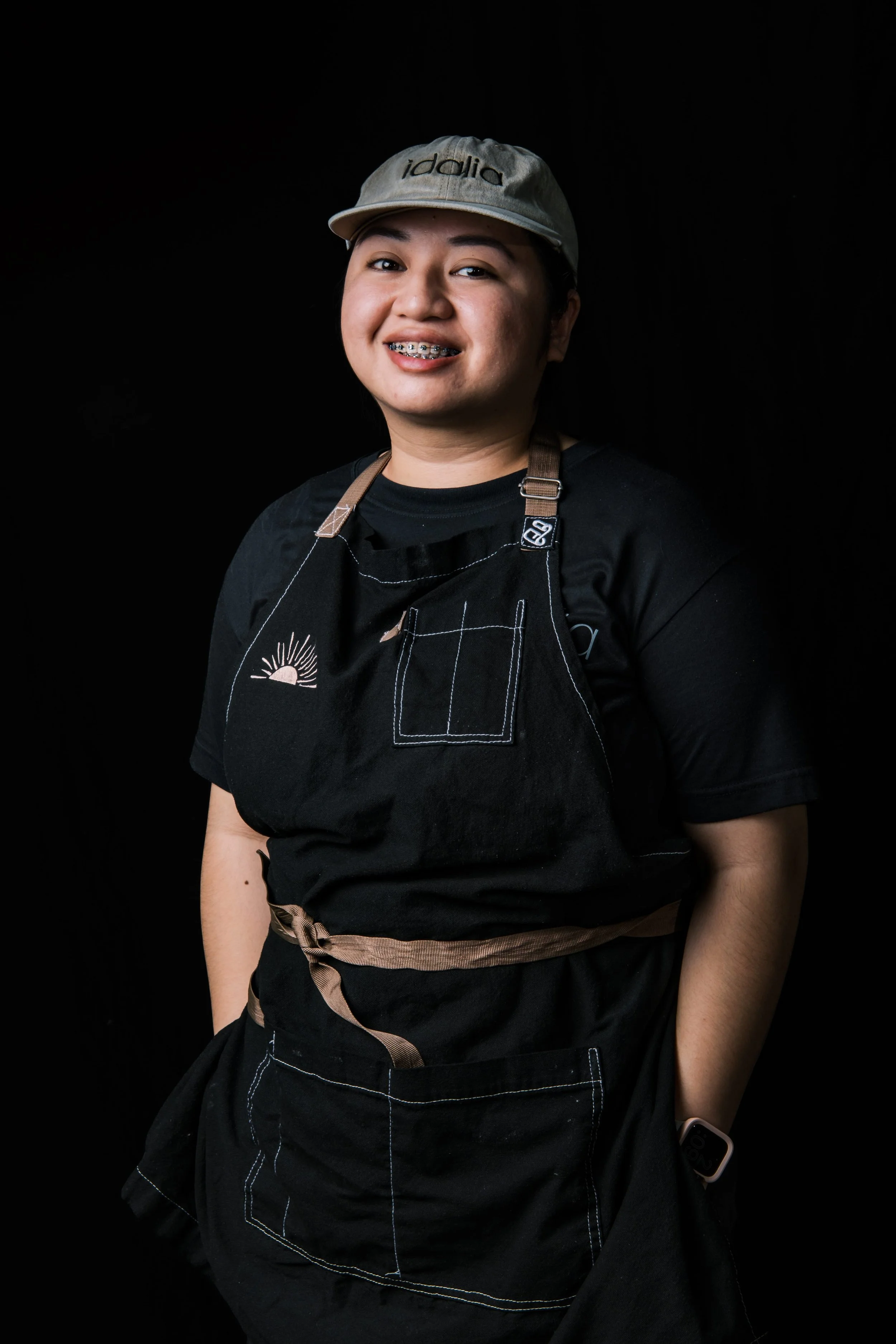 Aya Rances-Danao - Portrait of a young woman smiling, wearing a beige cap, black apron, and a black t-shirt, standing against a black background.