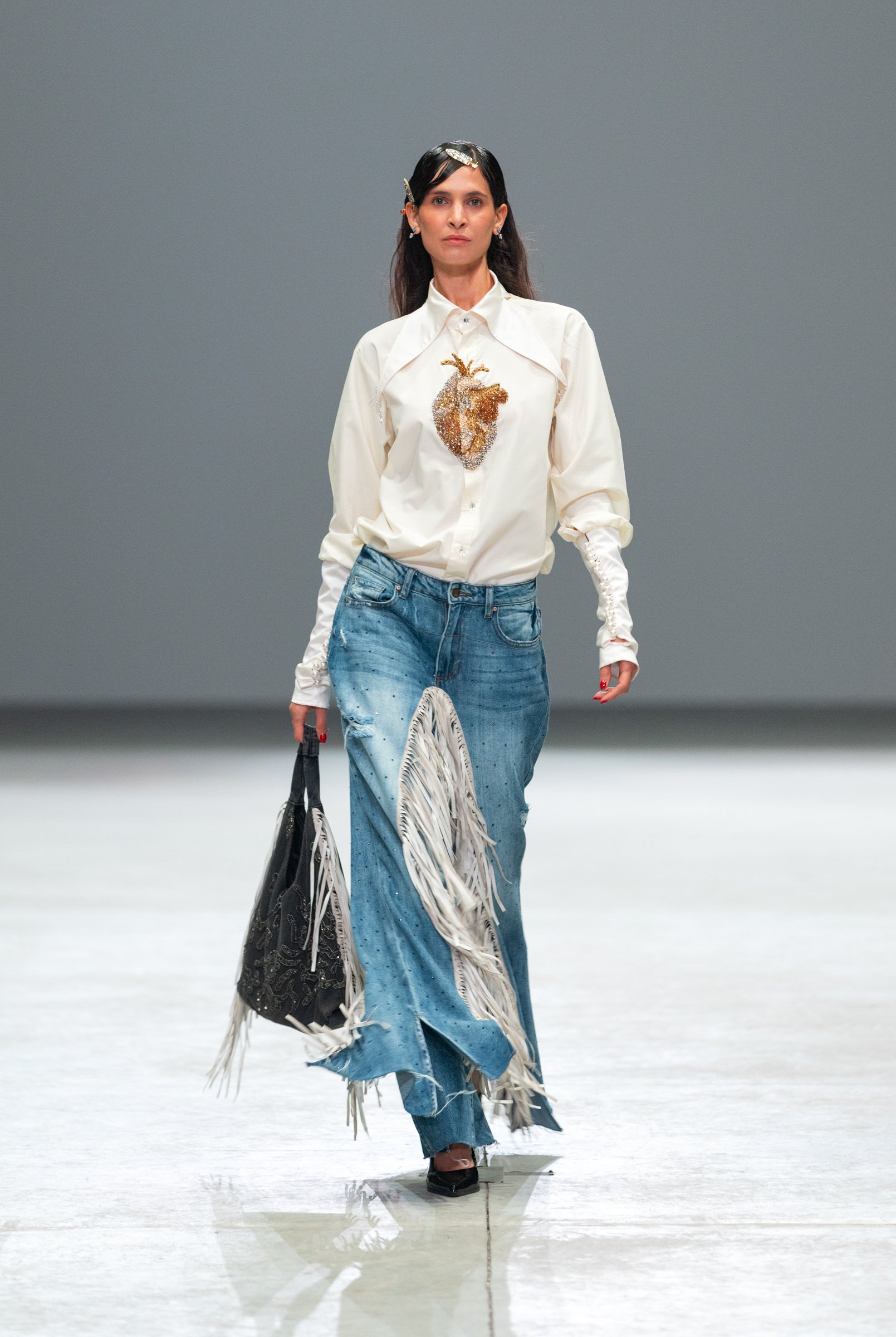  Model in statement denim jeans and a white blouse with a bold embroidered heart, part of Vero Solis’ collection at Volvo Fashion Week Mexico 2025. Emotionally charged, handcrafted, and proudly Mexican. 