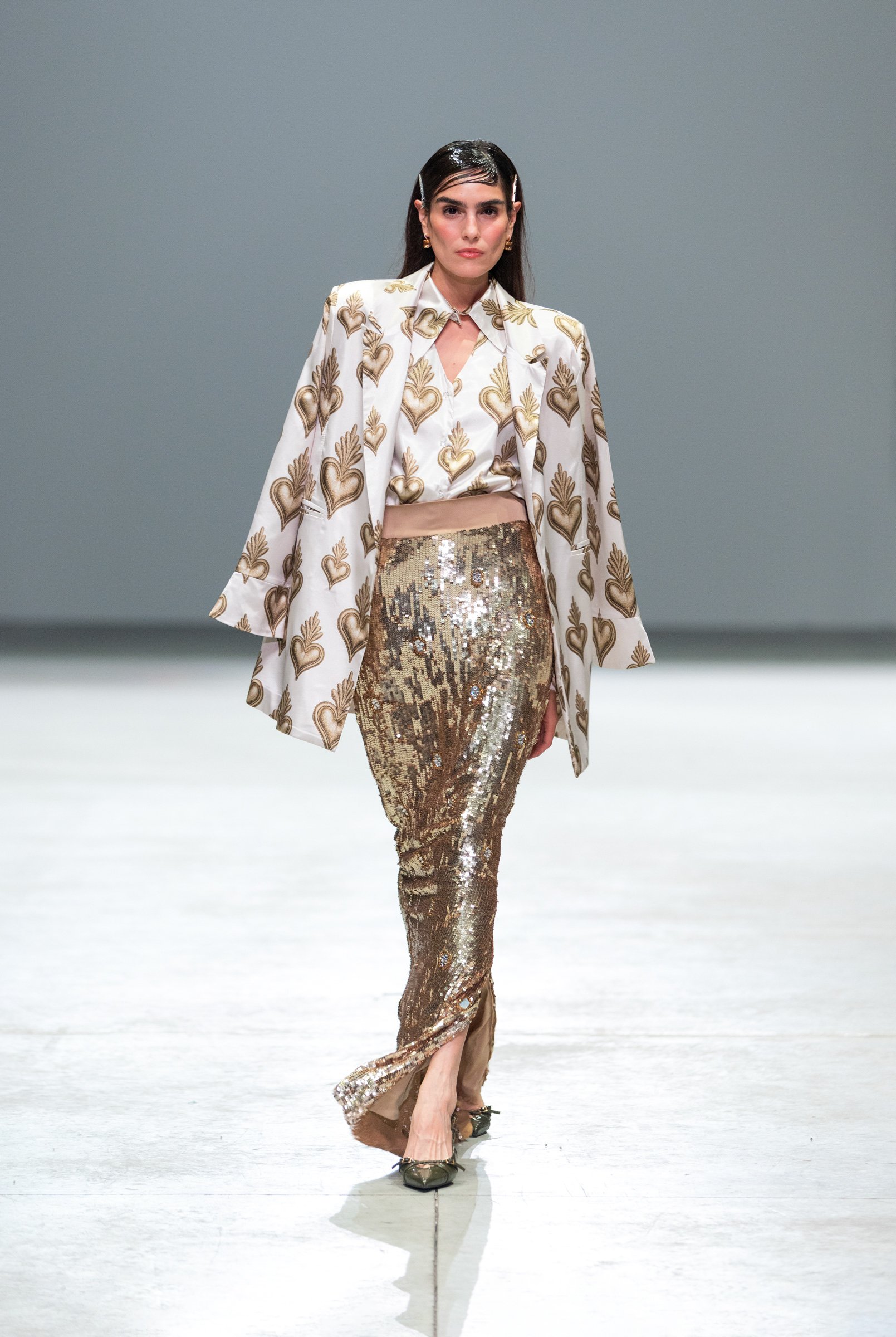  Glamorous runway shot of a model wearing a golden sequin dress and a refined white blazer with golden heart prints — the essence of Vero Solis’ vibrant elegance captured by KNYVAL. 