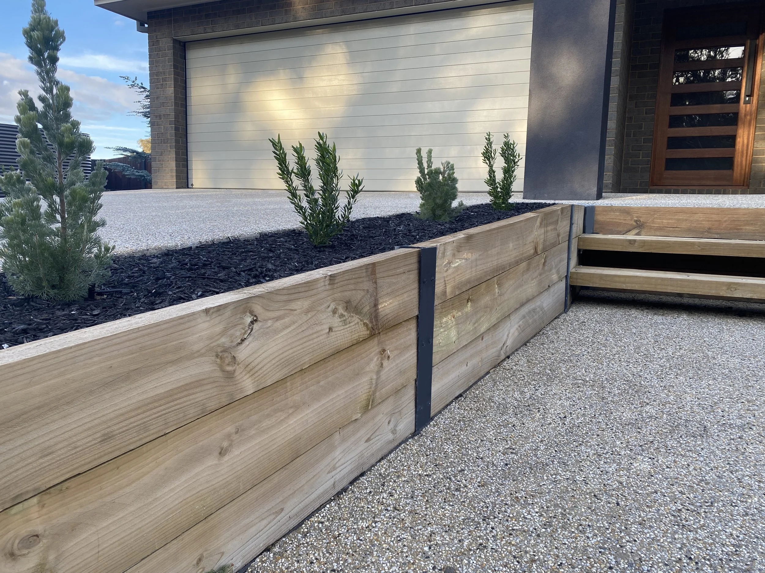 A wooden raised garden bed with small green plants, black metal corner connectors, and gravel walkway in front of a modern house with garage door and front door.