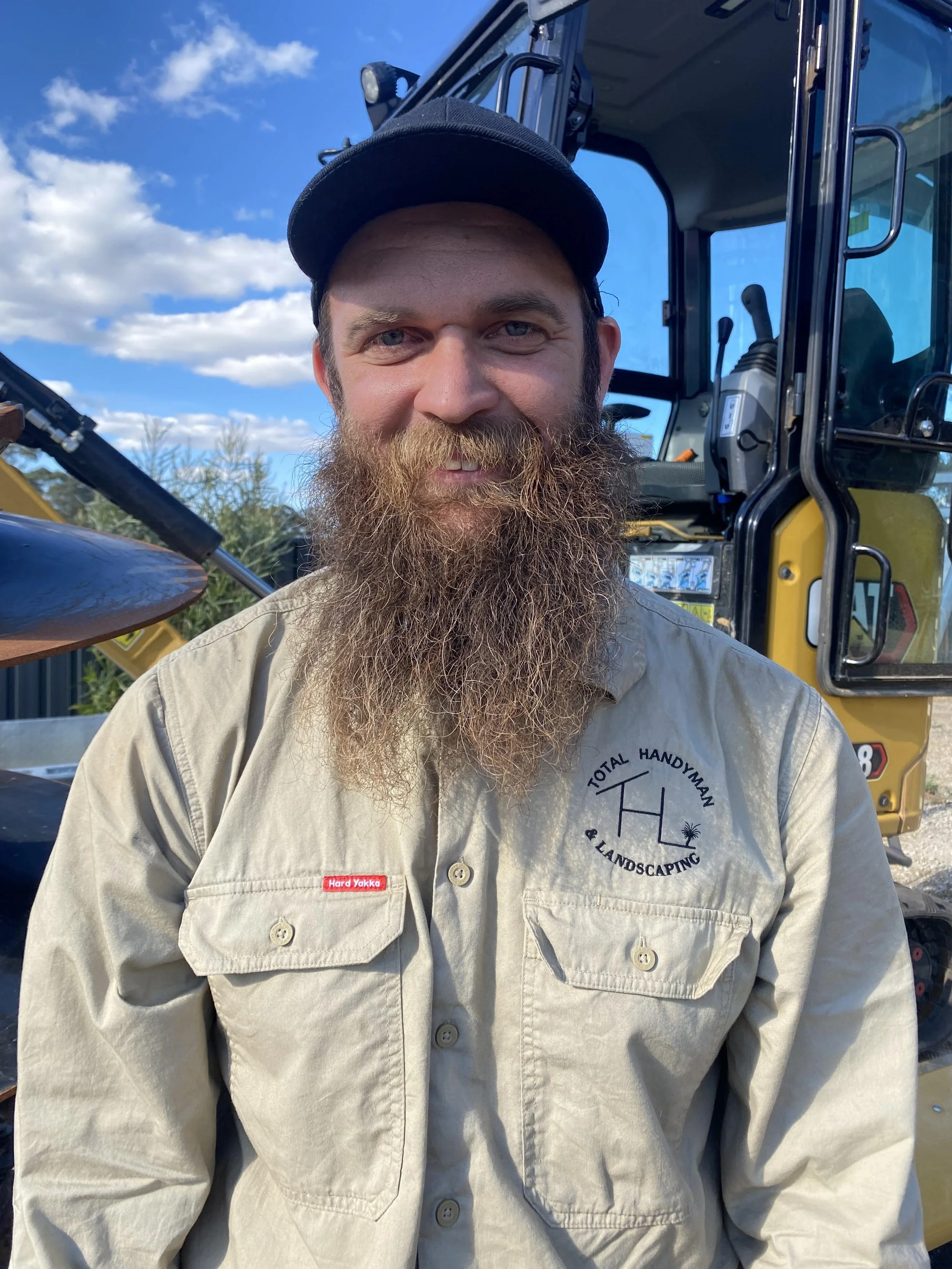A man with a long beard, wearing a black cap and beige work shirt with a logo and red tag, standing in front of construction equipment outdoors on a partly cloudy day. The logo says Total Handyman and Landscaping.