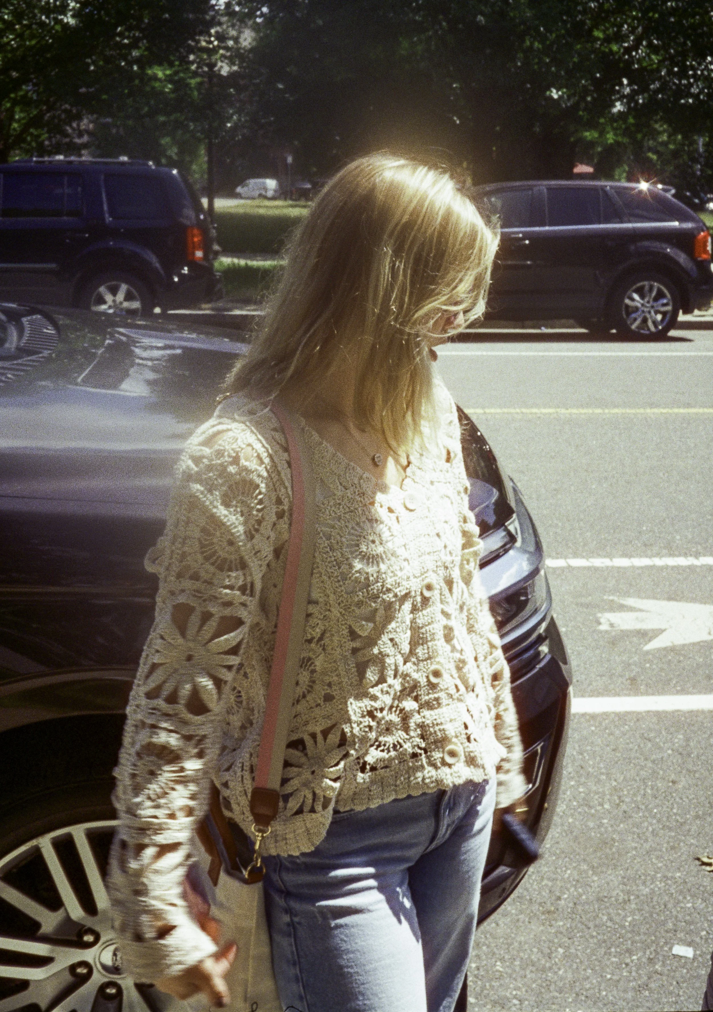 A woman with shoulder-length blonde hair wearing a beige crocheted sweater and light blue jeans, standing outdoors near a car on a sunny day.