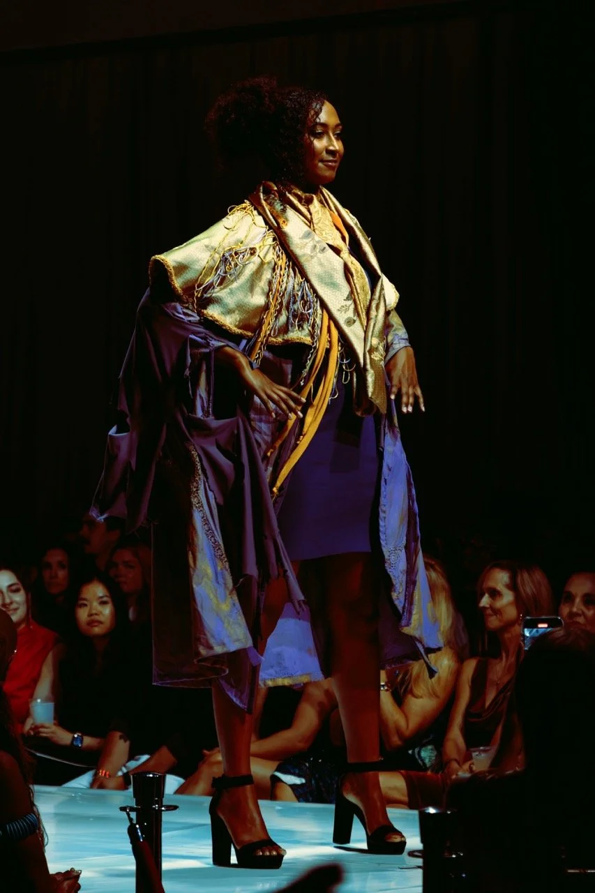 A woman walking on a runway during a fashion show, wearing high heels and a colorful, layered outfit with a patterned shawl or jacket, with an audience watching in the background.