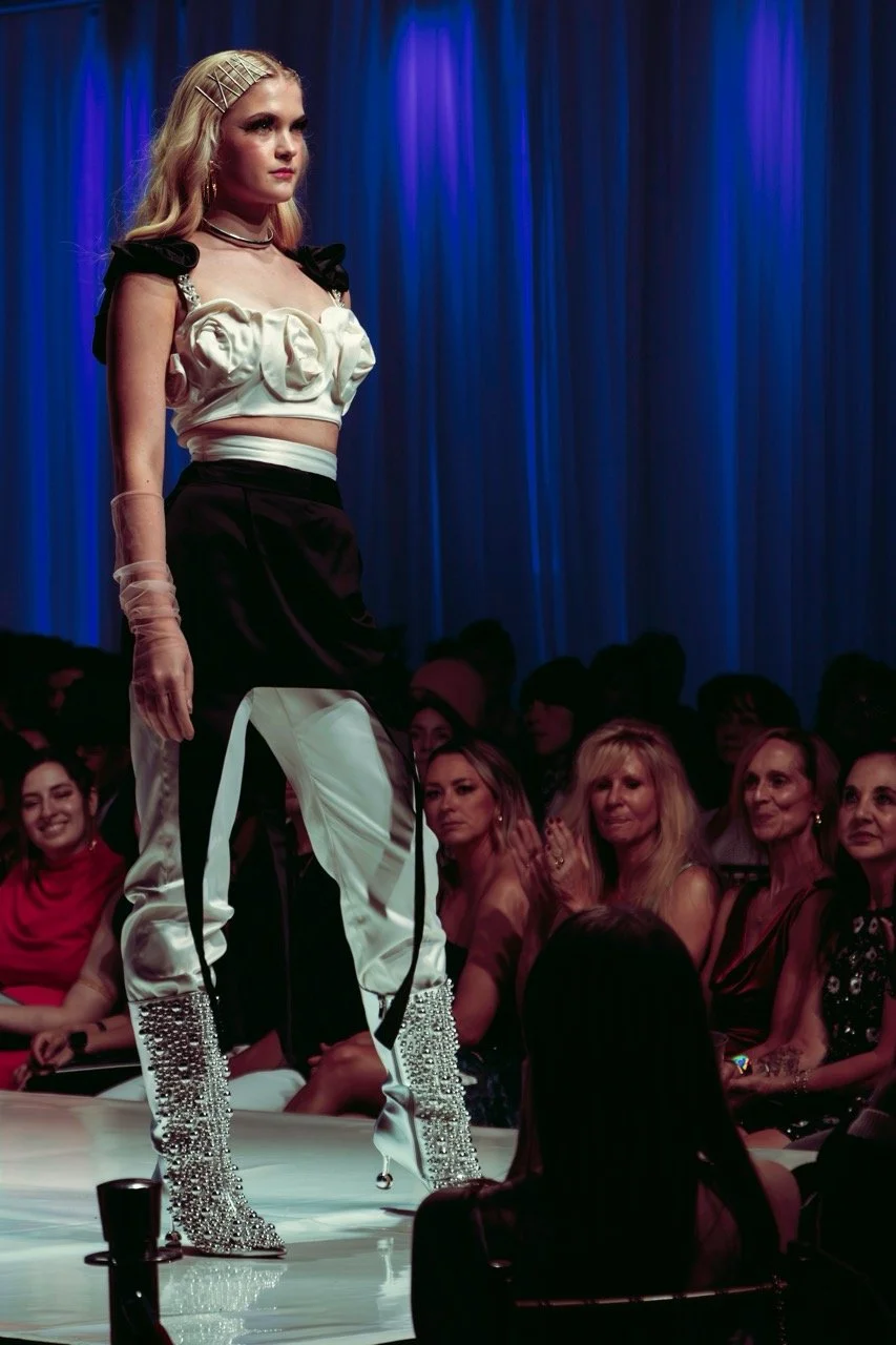 A fashion runway model walking on stage in front of an audience, wearing a stylish outfit with pearl embellishments and rhinestone boots, under blue stage lighting.