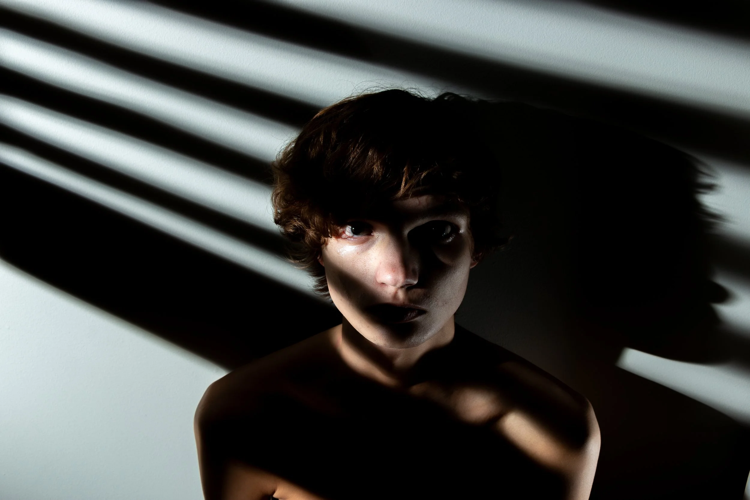 A person with short, curly hair is partially illuminated by sunlight through window blinds, casting dramatic shadows on their face and upper body.