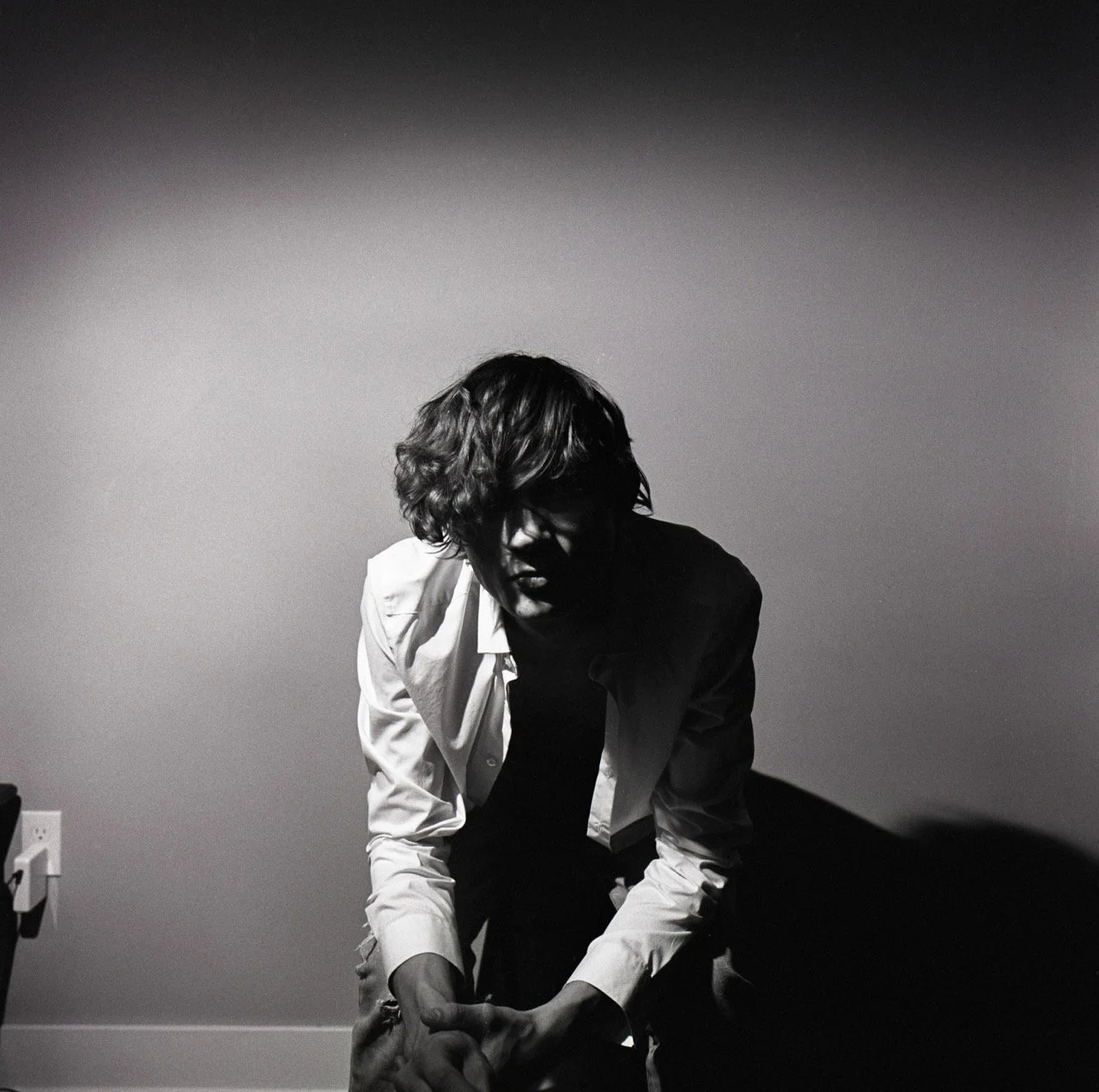 A black and white photograph of a person with wavy hair, wearing a white shirt over a dark top, sitting or crouching against a plain wall with minimal shadows.