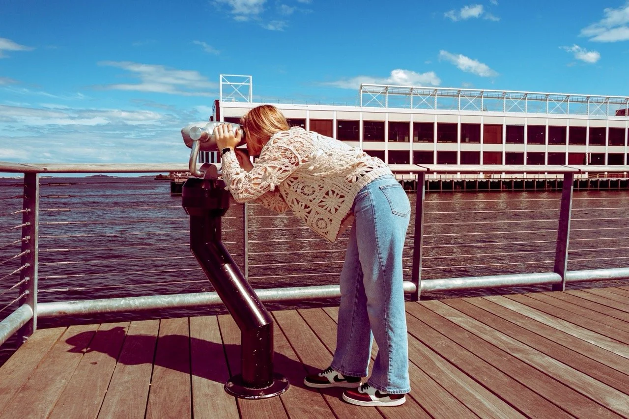 A woman with red hair, wearing a beige lace top and blue jeans, looking through a coin-operated binocular viewer on a wooden pier by a river or lake during the day with a partly cloudy sky.