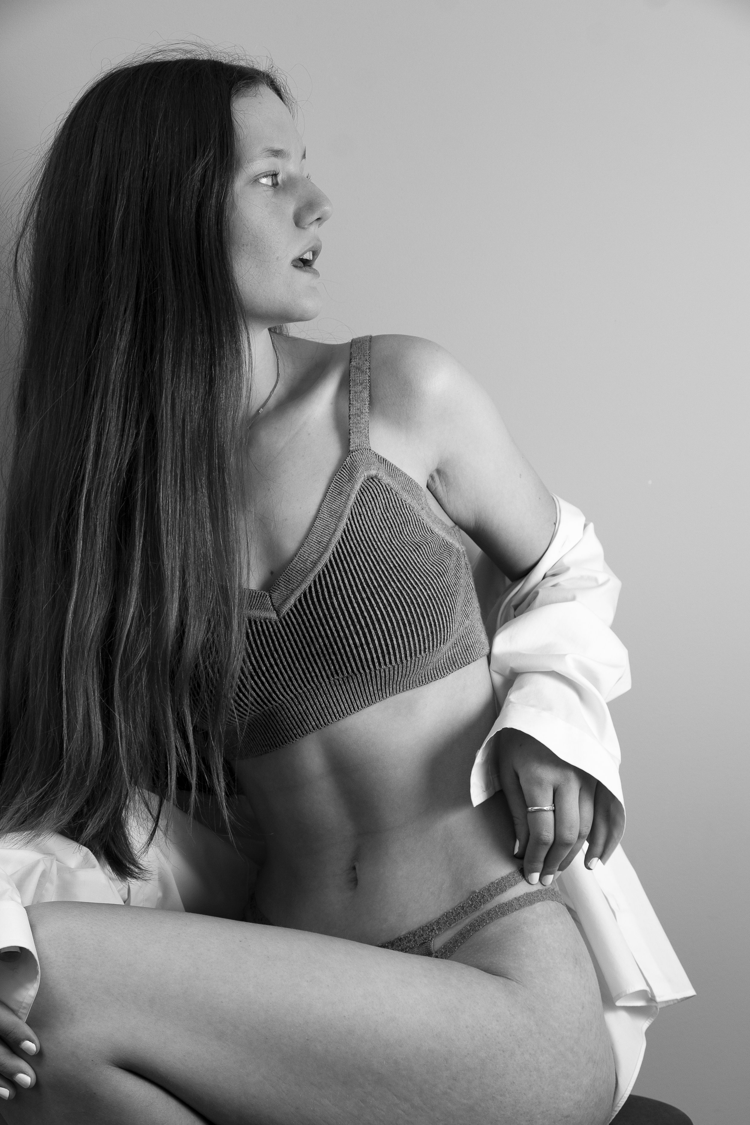 A woman with long hair looking to the left, wearing underwear and an open shirt, sitting against a plain background.