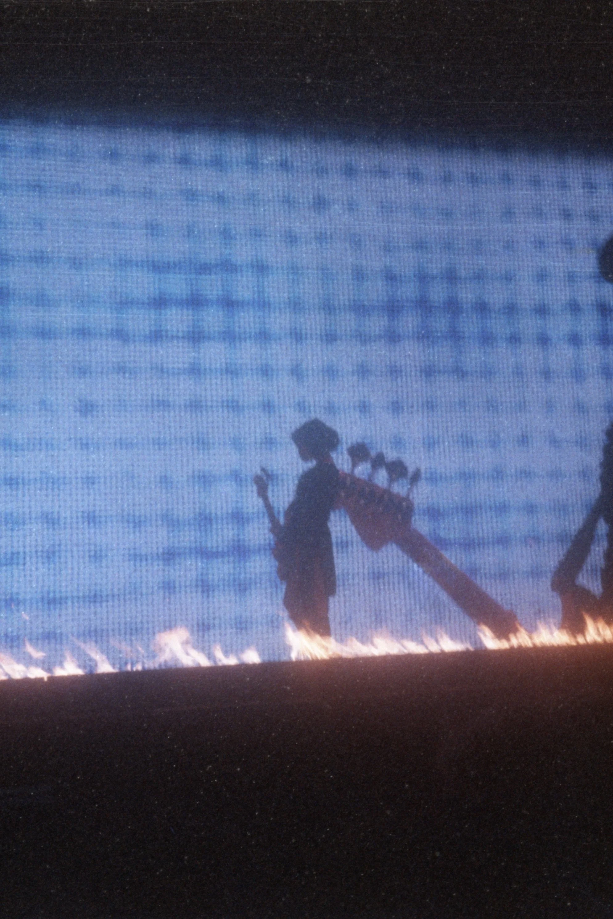 Silhouette of a person holding a guitar, walking near a row of fire on the ground, with a blue background.