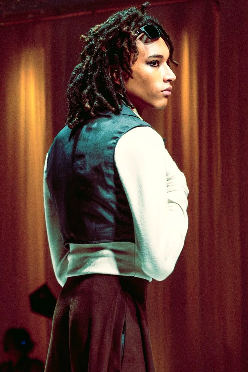 A woman with dark curly dreadlocks and sunglasses resting on her head, standing to the side with her arms crossed, wearing a teal vest over a white long-sleeve shirt, indoors with wooden paneling in the background.