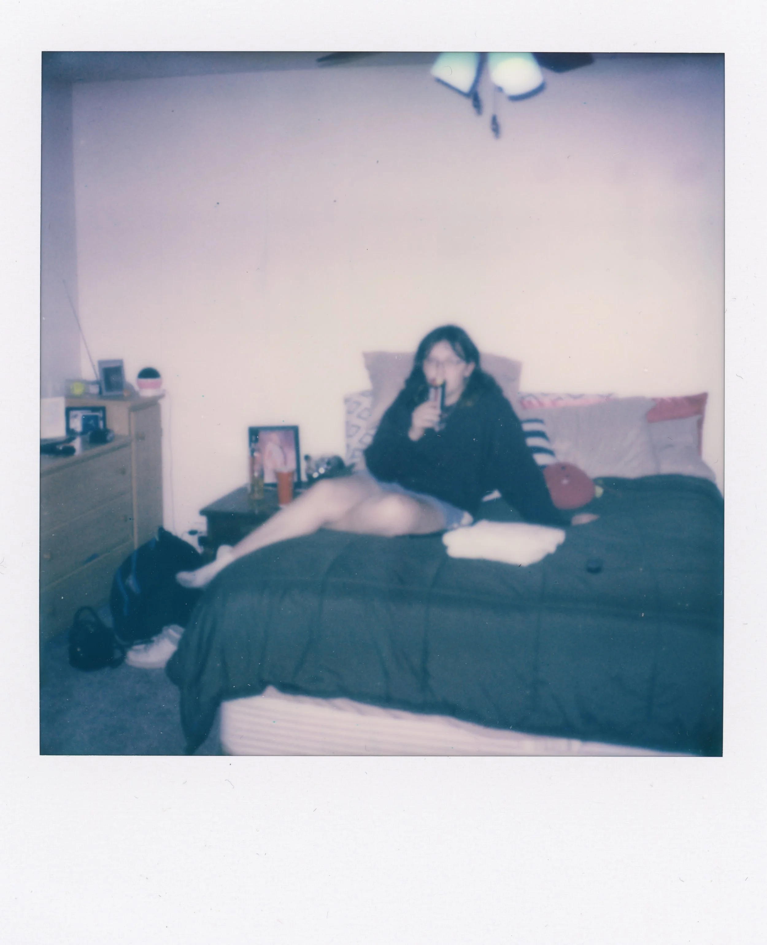 A woman sitting on a bed drinking from a glass in a bedroom with a nightstand, dresser, and framed photo.