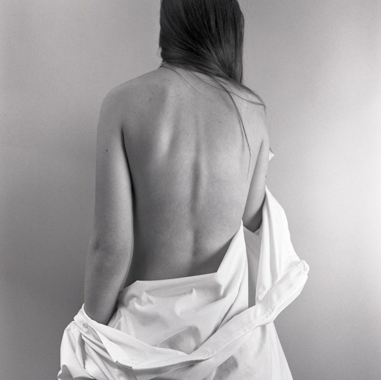 Black and white photograph of a woman with her back to the camera, partially covered with a white sheet, revealing her back and shoulder.