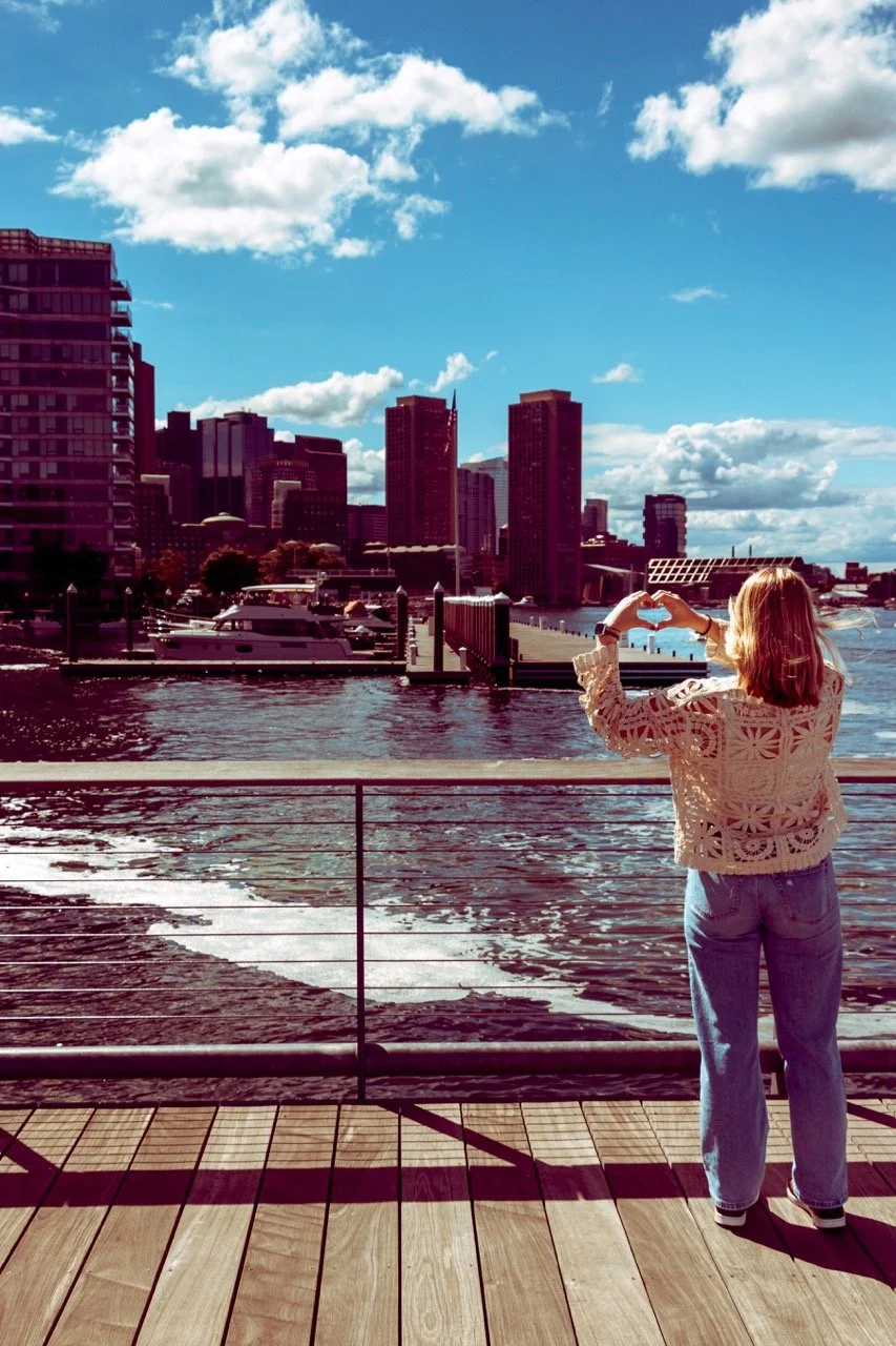 A woman standing on a wooden pier by the water, making a heart shape with her hands, facing a city skyline with tall buildings, boats docked, and a partly cloudy sky.