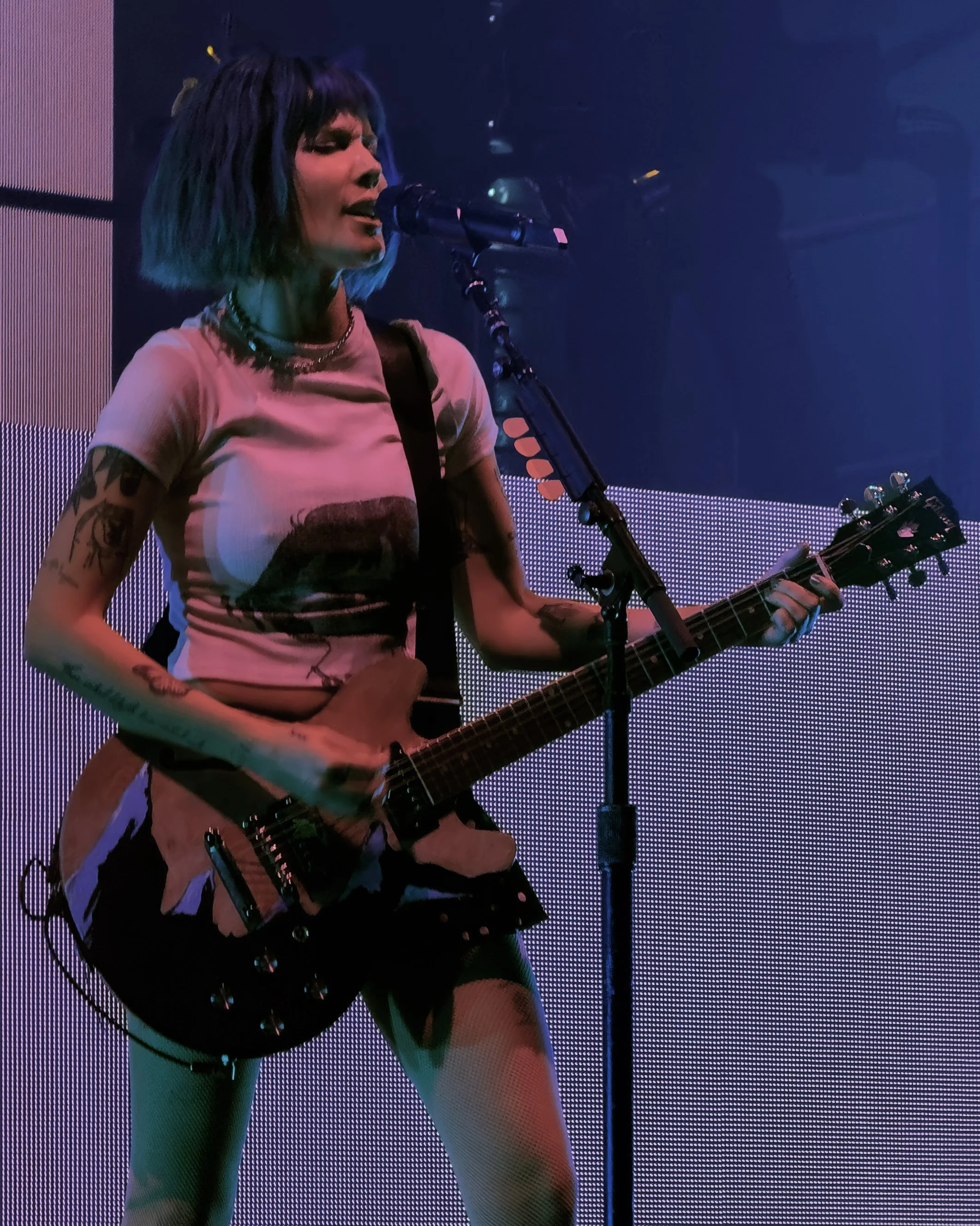 A woman with short, blue-tipped hair singing and playing an electric guitar on stage with colorful lighting.
