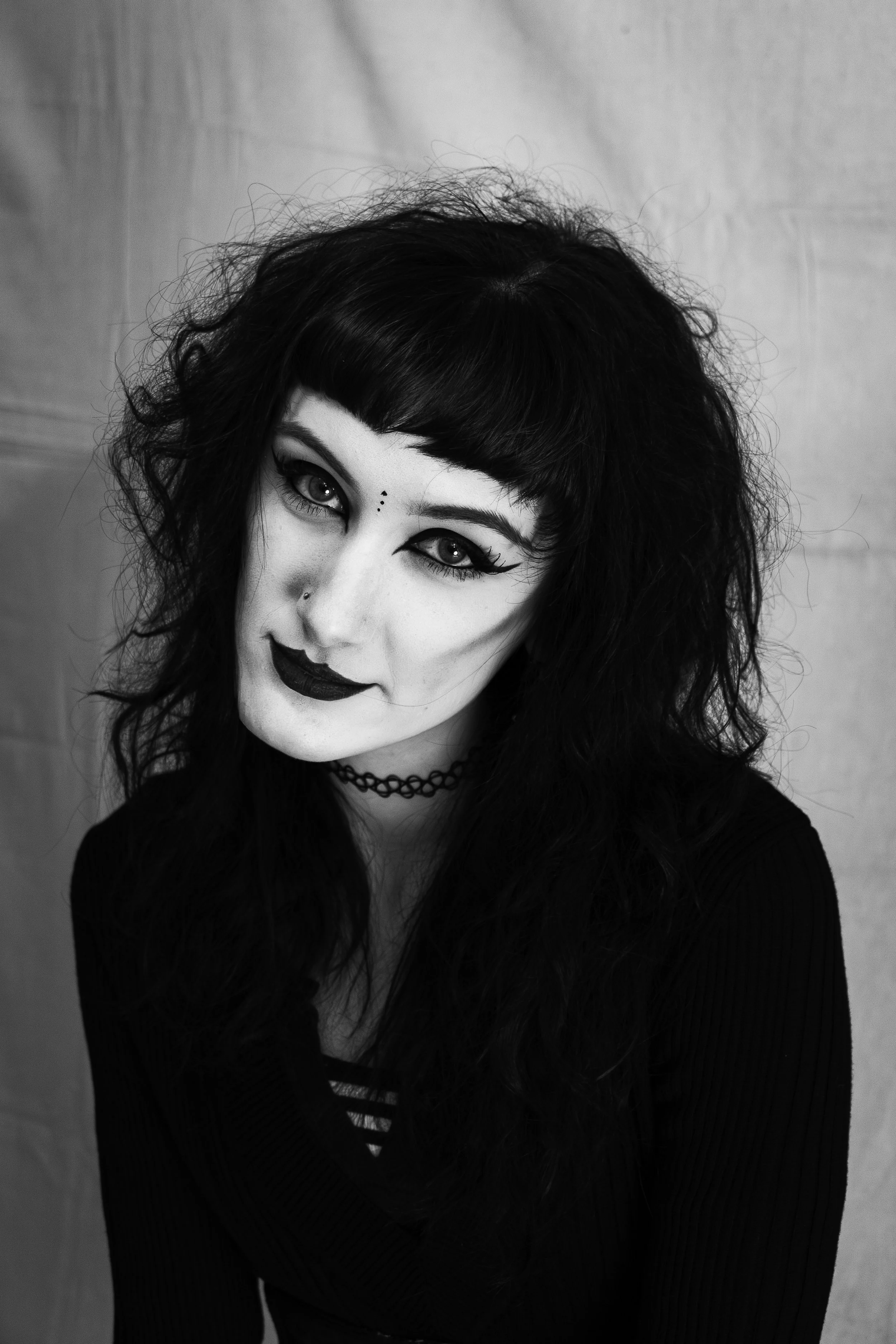 Black and white portrait of a woman with long, curly black hair, dark makeup, and a choker necklace, looking at the camera.