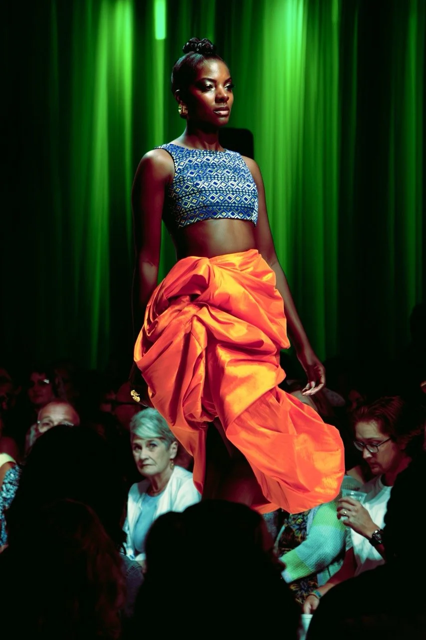 A runway model in a blue patterned crop top and bright orange skirt walking on stage in front of an audience with green curtains in the background.