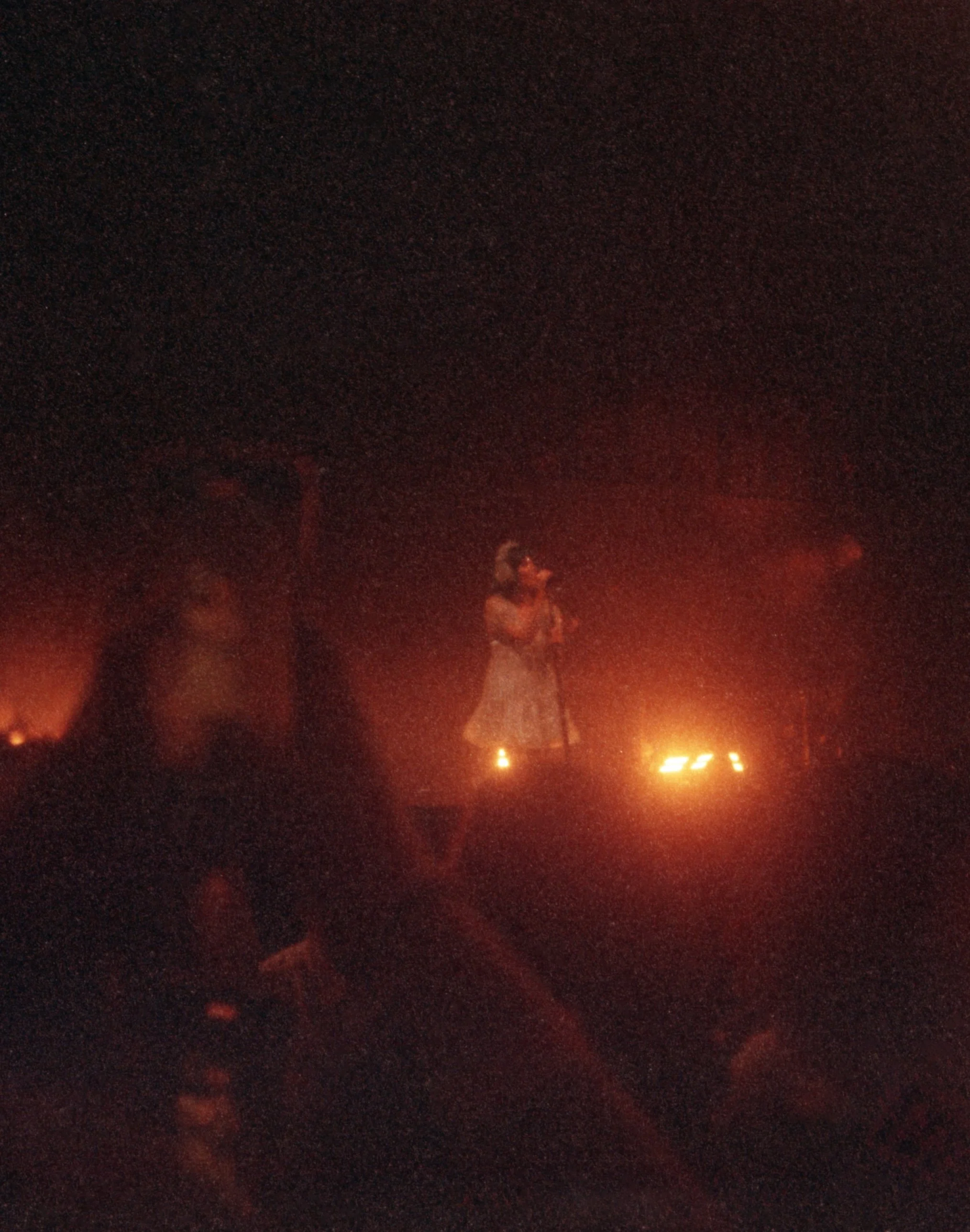A singer performing on stage at night, illuminated by small lights or candles, with audience members visible in the dark.