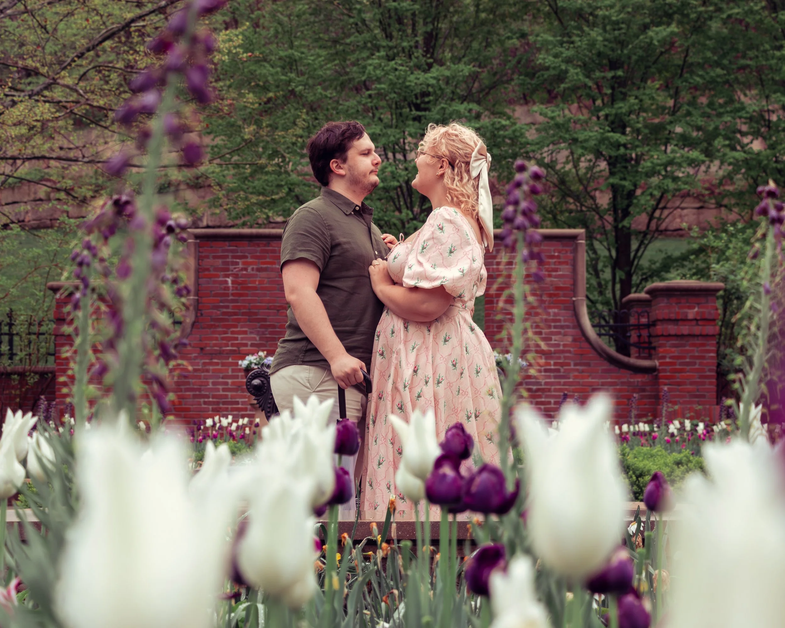 A couple stands close together in a garden with tulips, facing each other with their eyes locked. The man holds a suitcase and a phone in his hand, and the woman has a white bow in her curly blonde hair. In the background, there is a red brick wall a