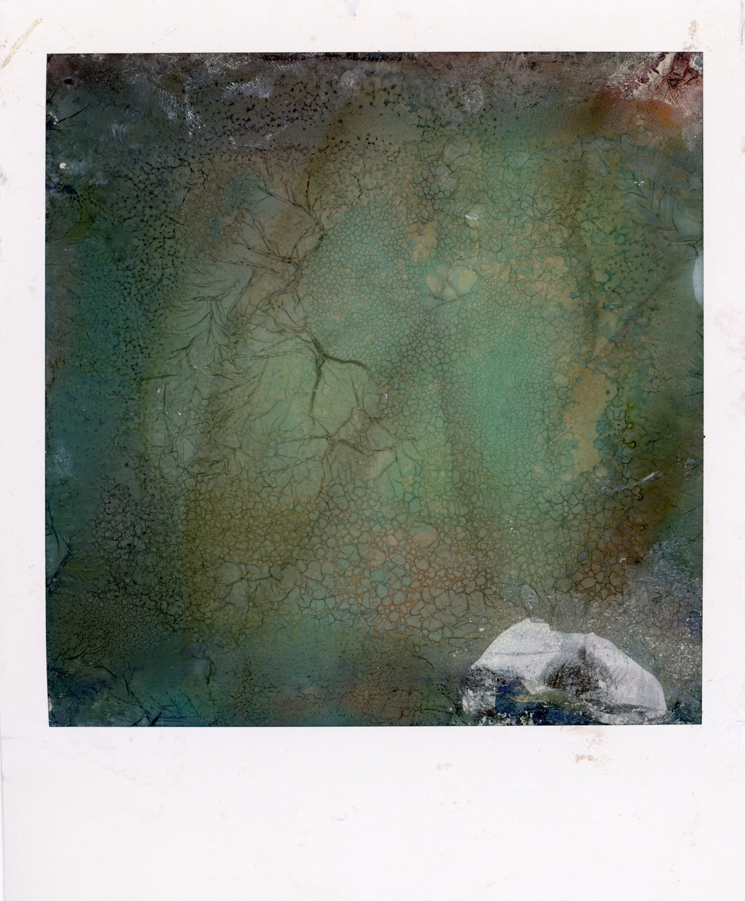 An abstract image with cracked, textured patterns in shades of green, brown, and blue, with a small white object partially visible at the bottom right corner.