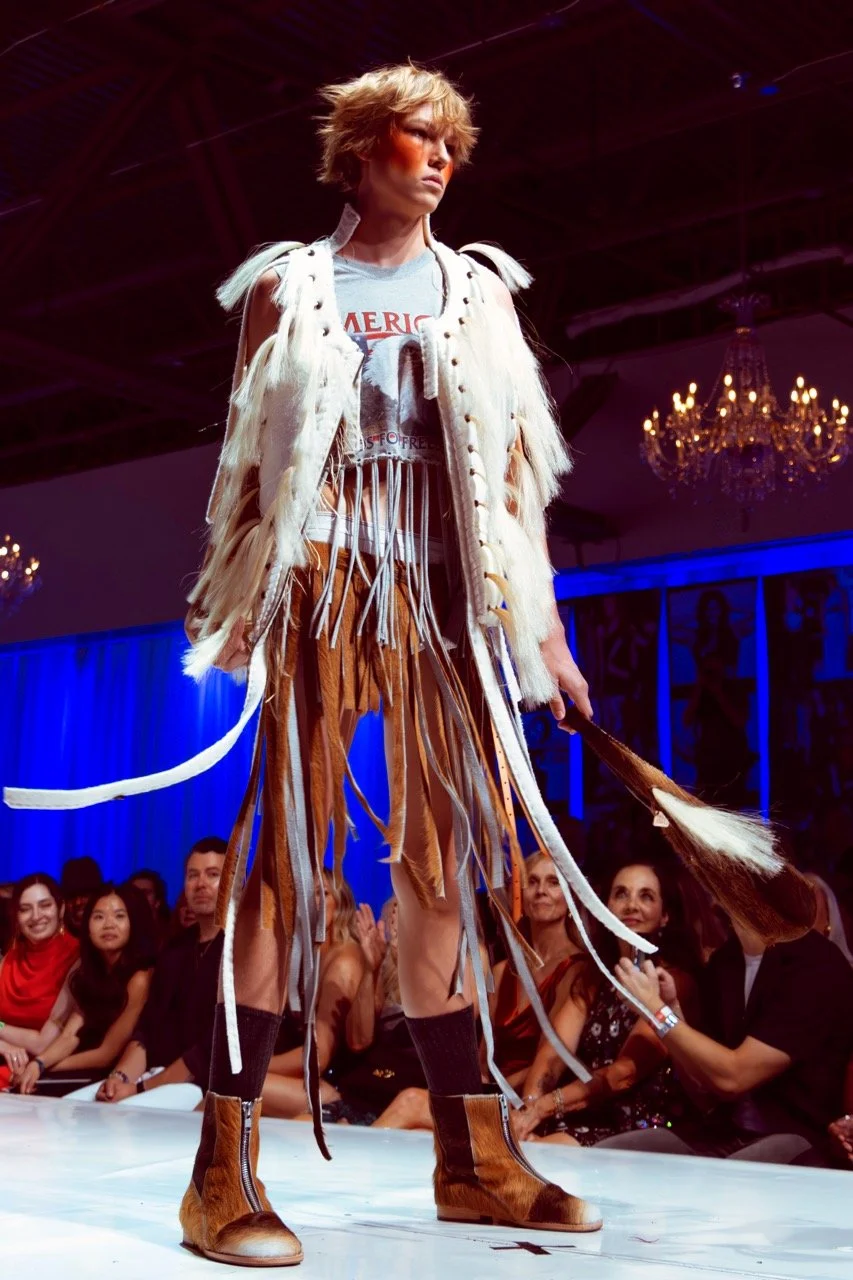 Model on runway wearing an eclectic outfit with a fringed graphic t-shirt, a fur vest, fringed pants, and cowboy boots, with audience watching in the background at fashion show.