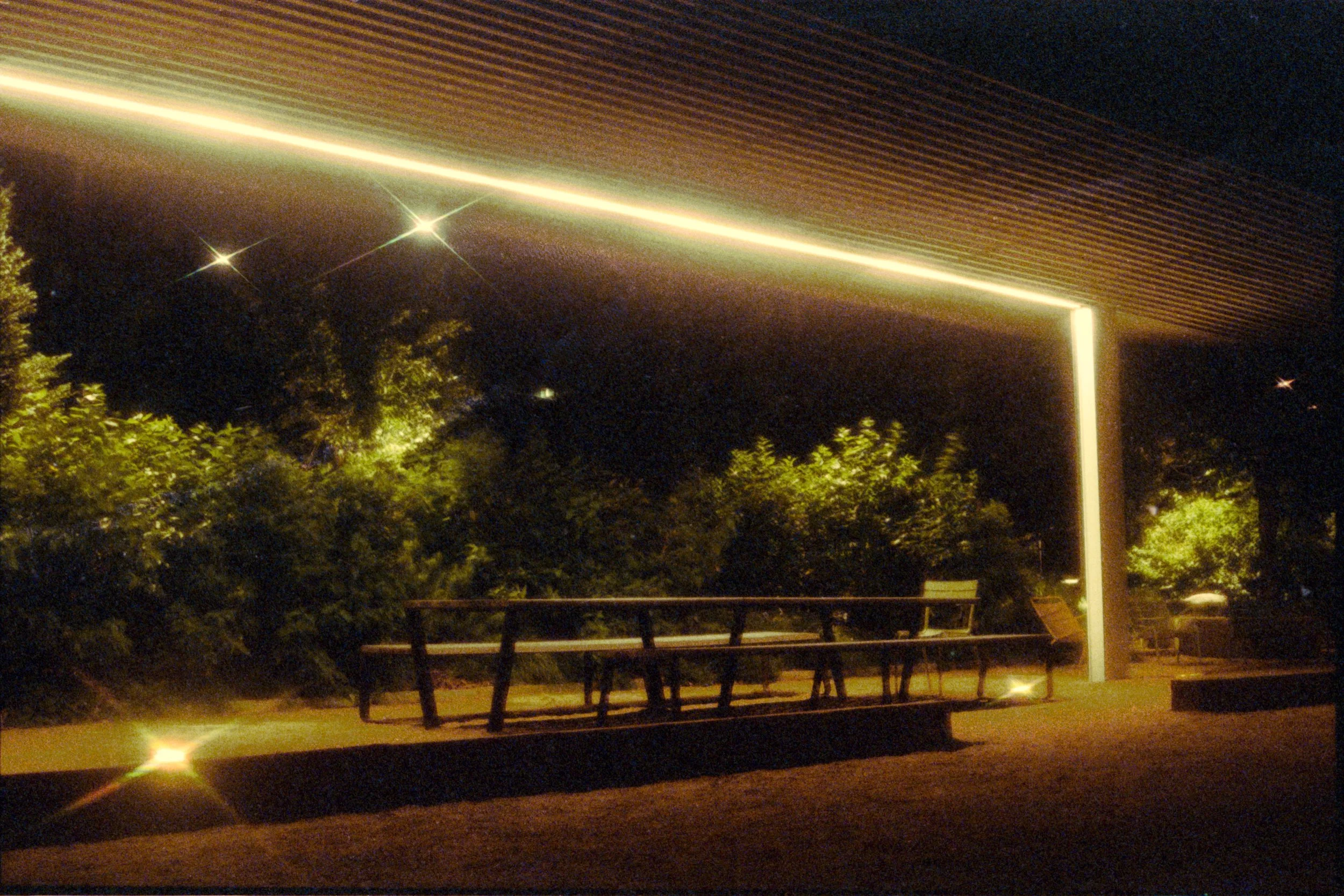 Nighttime scene of a covered outdoor seating area with a bench and chairs, illuminated by a bright strip of light on the ceiling and reflecting light on the ground, surrounded by trees and bushes.