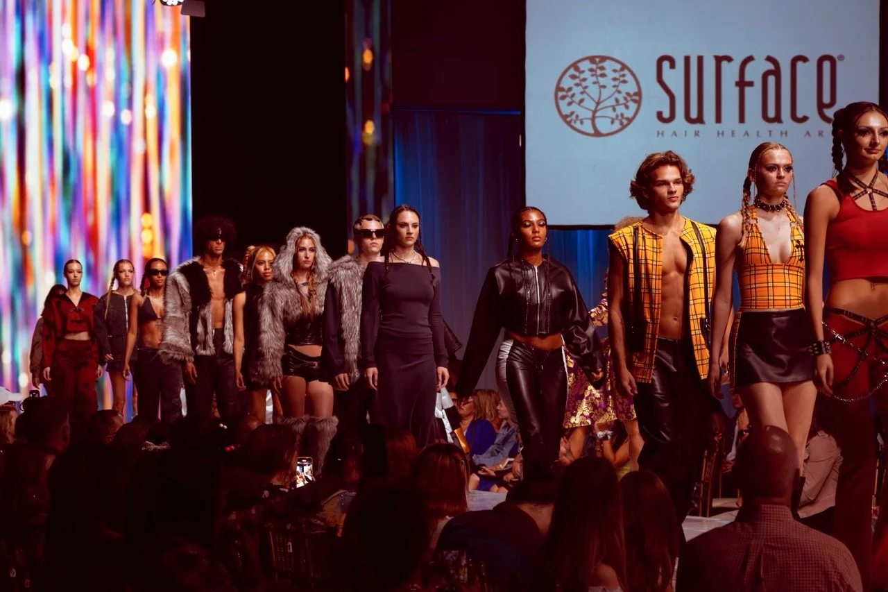 Models walking down runway during a fashion show with a large Surface Hair Health & Art logo projected on the screen behind them.