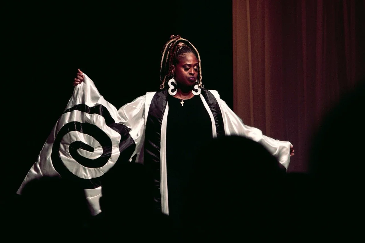 Woman on stage wearing a black dress and a white cape with black spiral design, also wearing large white earrings and a cross necklace, with a serious expression.