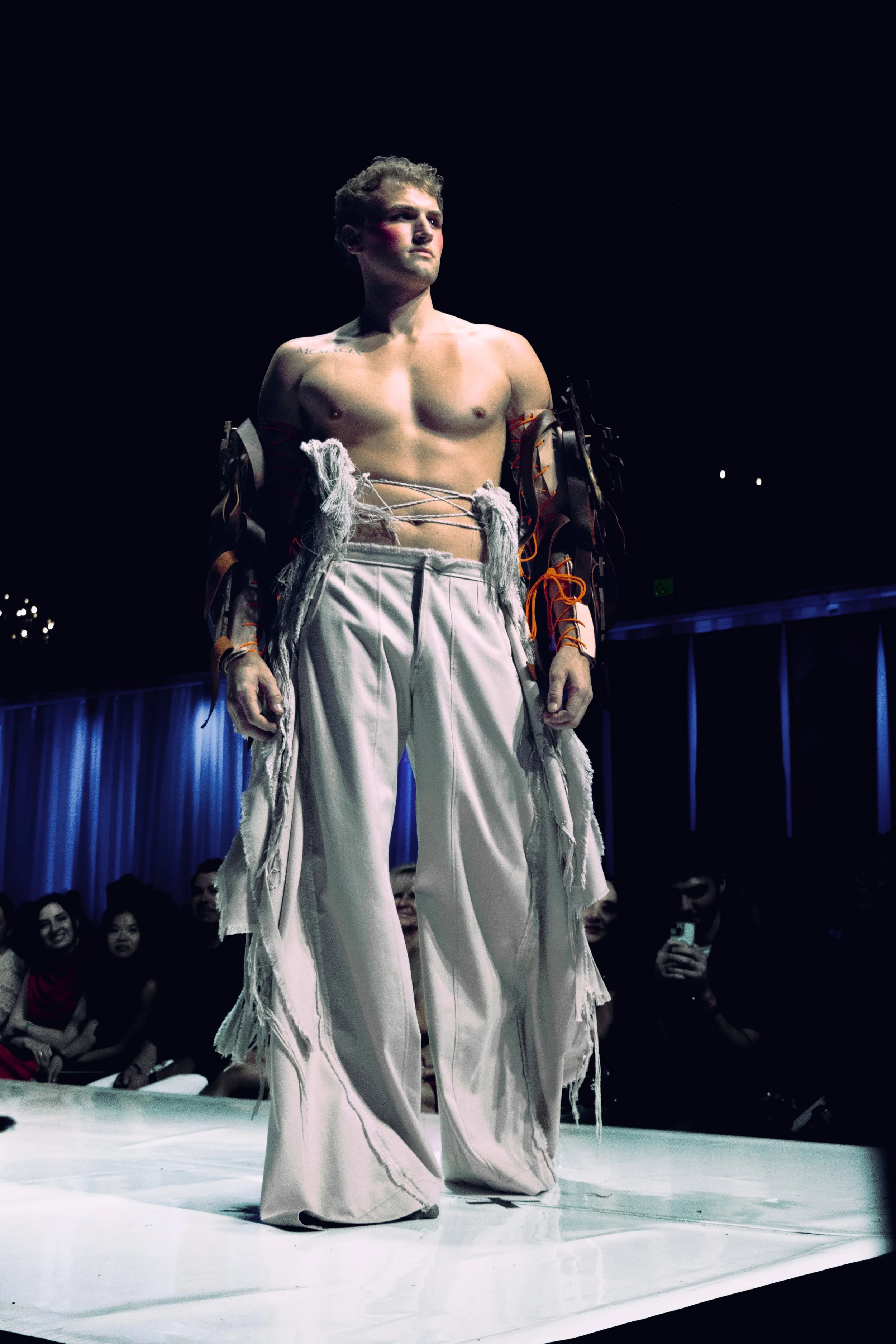 Male model walking on runway wearing distressed white pants and a unique, partially open top with fabric and string details, with audience watching in dimly lit fashion show setting.