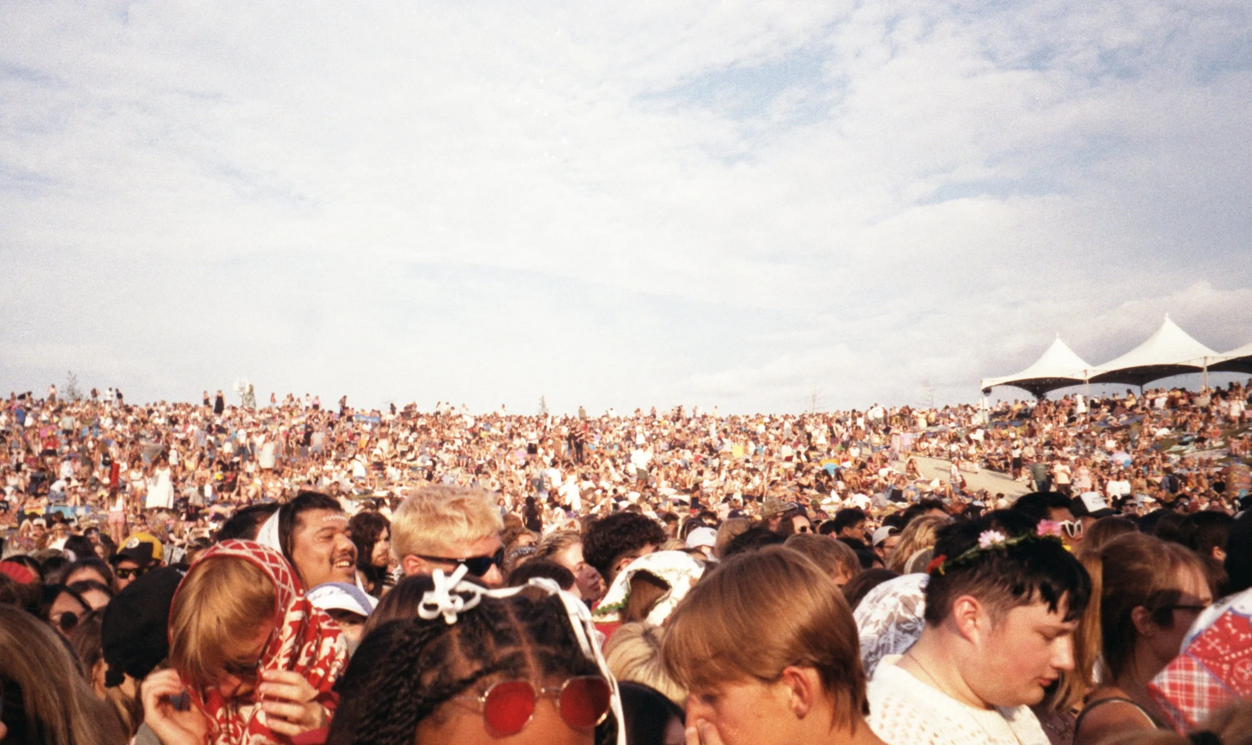 Crowd of people attending an outdoor event on a sunny day with tents in the background.