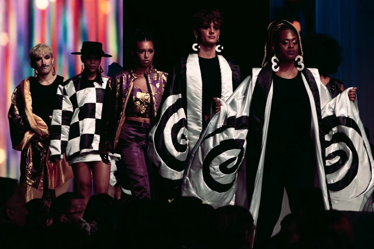 Fashion show runway with models wearing patterned and colorful outfits, including checkered coats and shiny fabrics, walking down the runway.