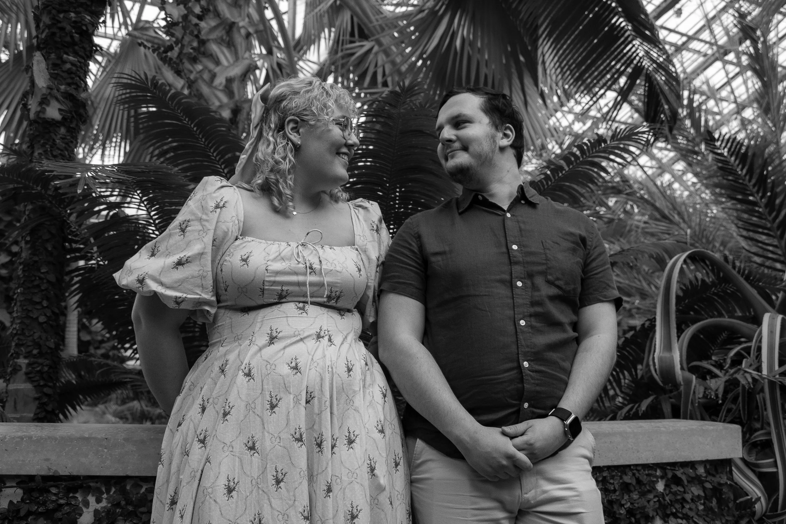 A black and white photo of a woman and a man standing close together outdoors, surrounded by tropical plants. The woman is smiling at the man, who is looking back at her. The woman wears a patterned dress with puffed sleeves, and the man wears a shor
