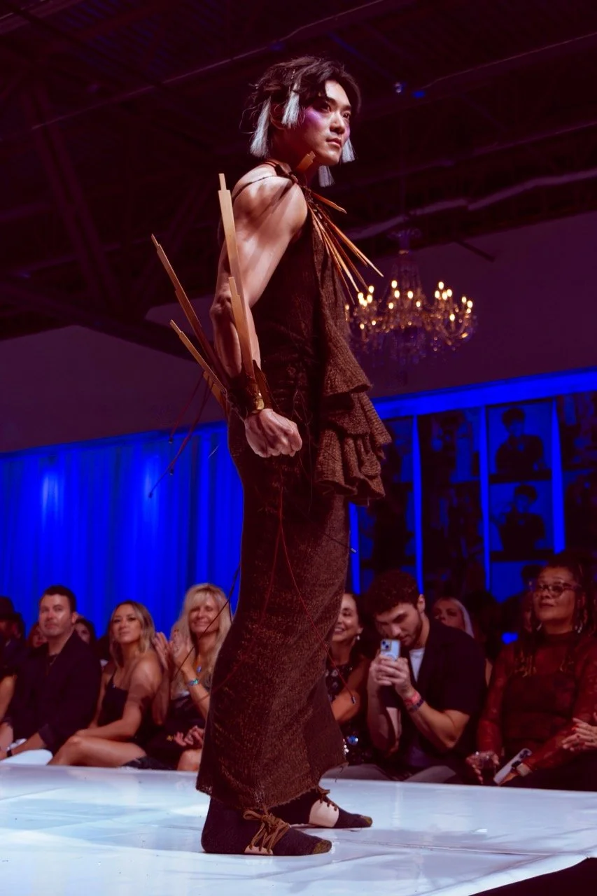 A model walking on a runway at a fashion show, wearing a brown textured outfit with large, sculptural wooden accessories on the arm and shoulder, with an audience watching and taking photos in a dimly lit room with chandeliers and blue lighting.