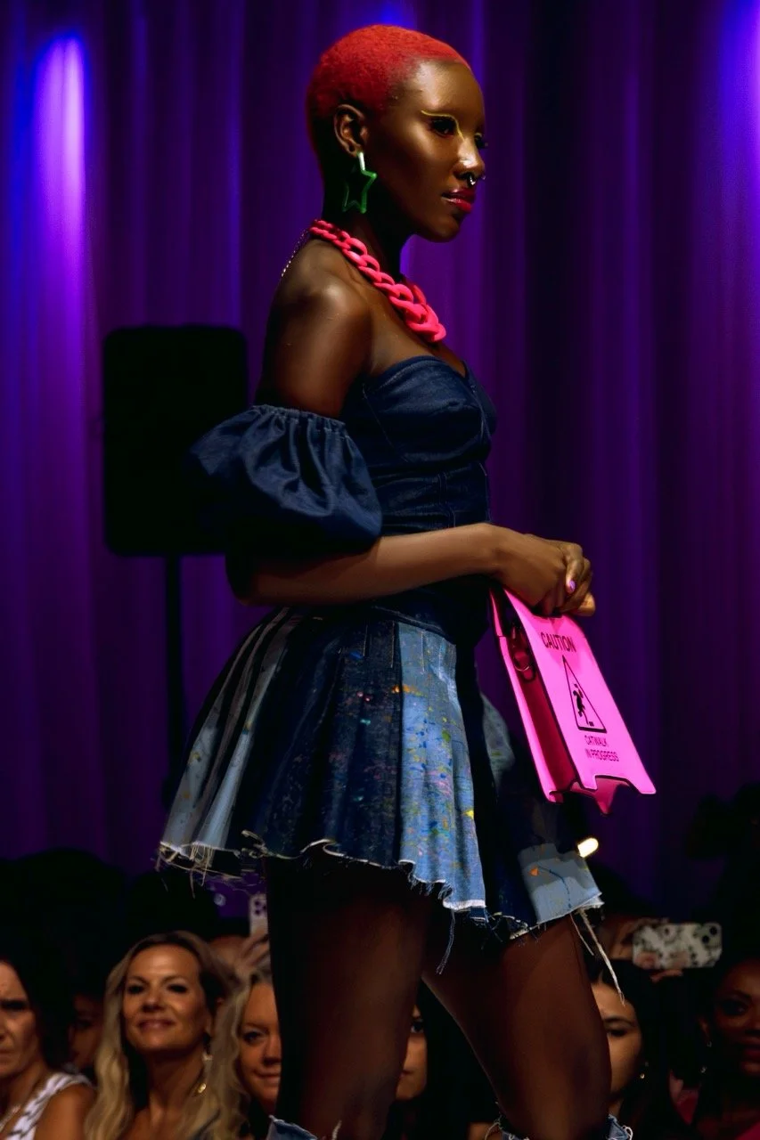 A woman with bright red hair on stage, wearing a dark off-the-shoulder top, a colorful denim skirt, large green star-shaped earrings, a chunky pink necklace, and holding a caution sign, with a purple curtain and an audience in the background.